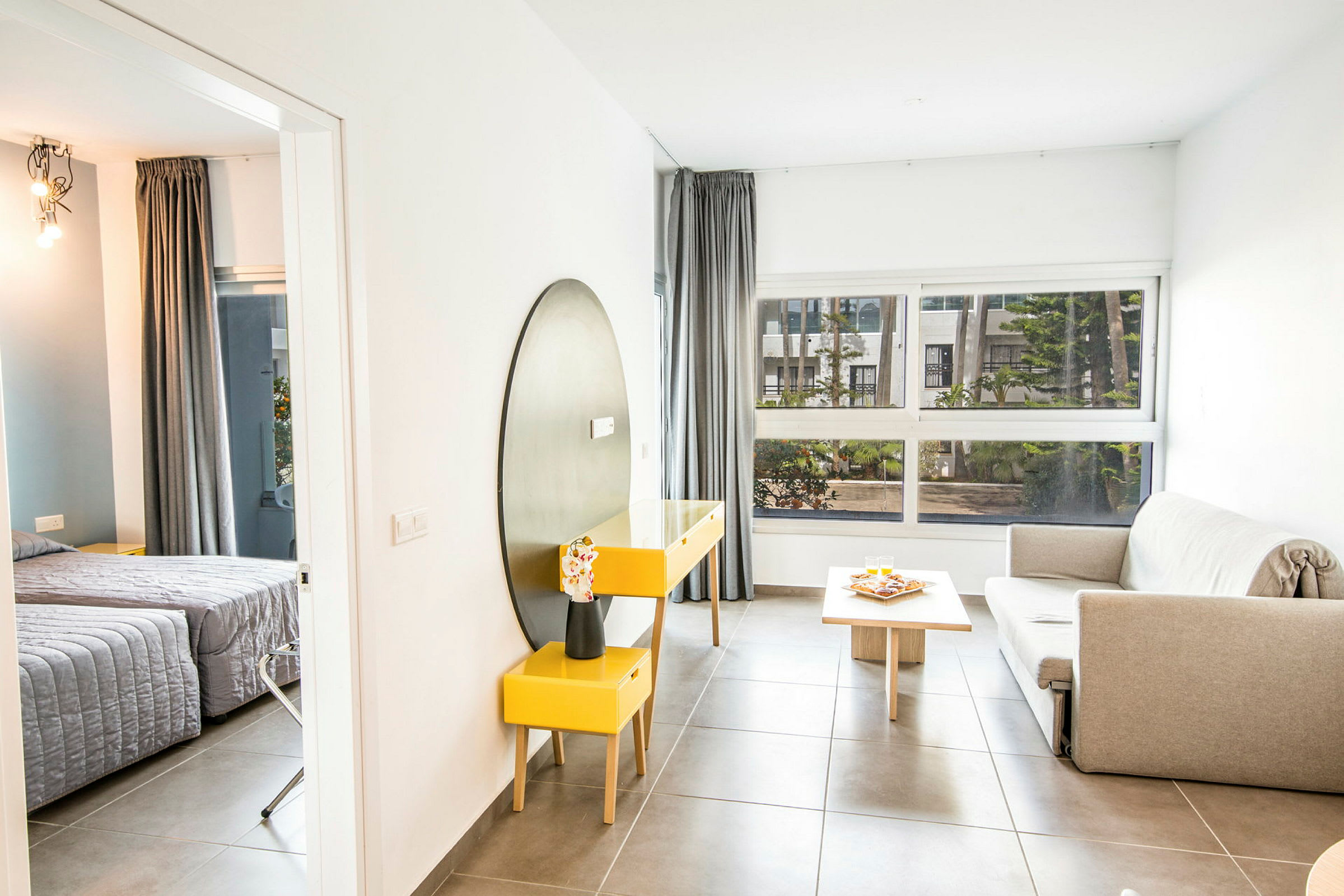 Ett modernt rum med soffhörna, bord och en stor fönstervy som släpper in naturligt ljus. I bakgrunden syns en dörröppning till sovrummet. (AI-genererad text)