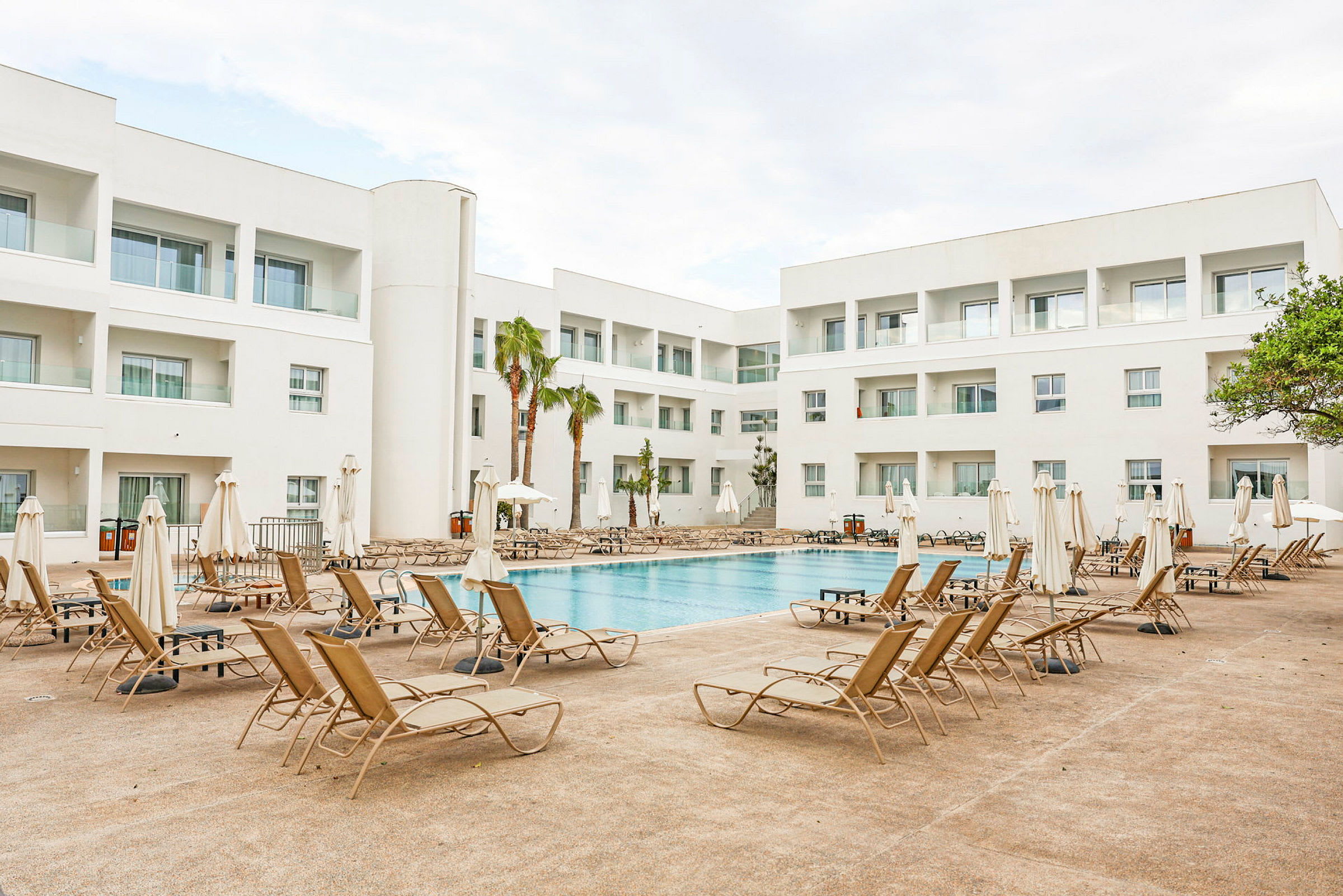 Utomhuspool omgiven av solstolar och vita byggnader i en hotellmiljö. (AI-genererad text)