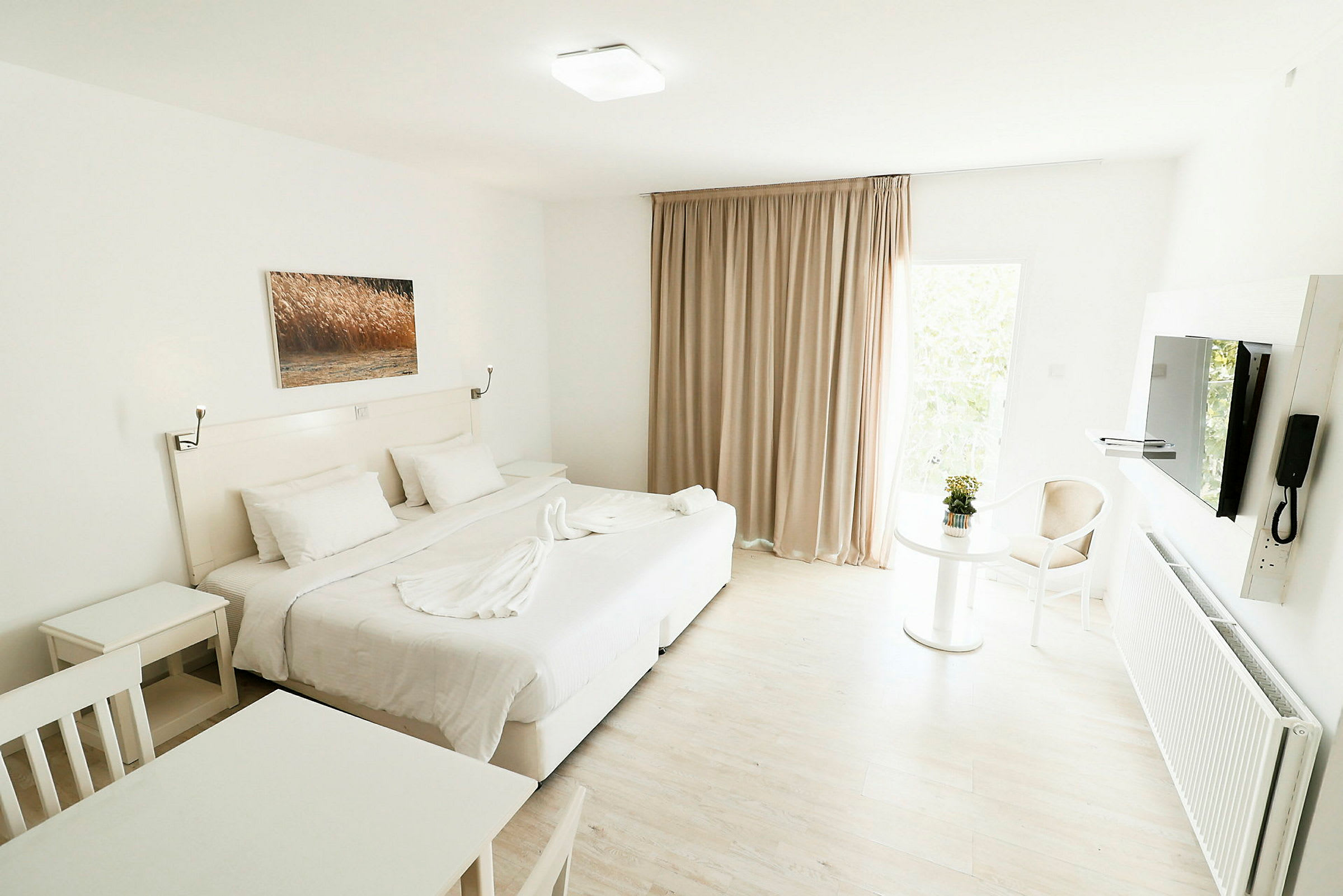 Ett ljust och modernt hotellrum med en stor säng, vita möbler och en glastavla på väggen. Ett bord med en blomma och en stol syns vid ett fönster med beige gardiner. (AI-genererad text)