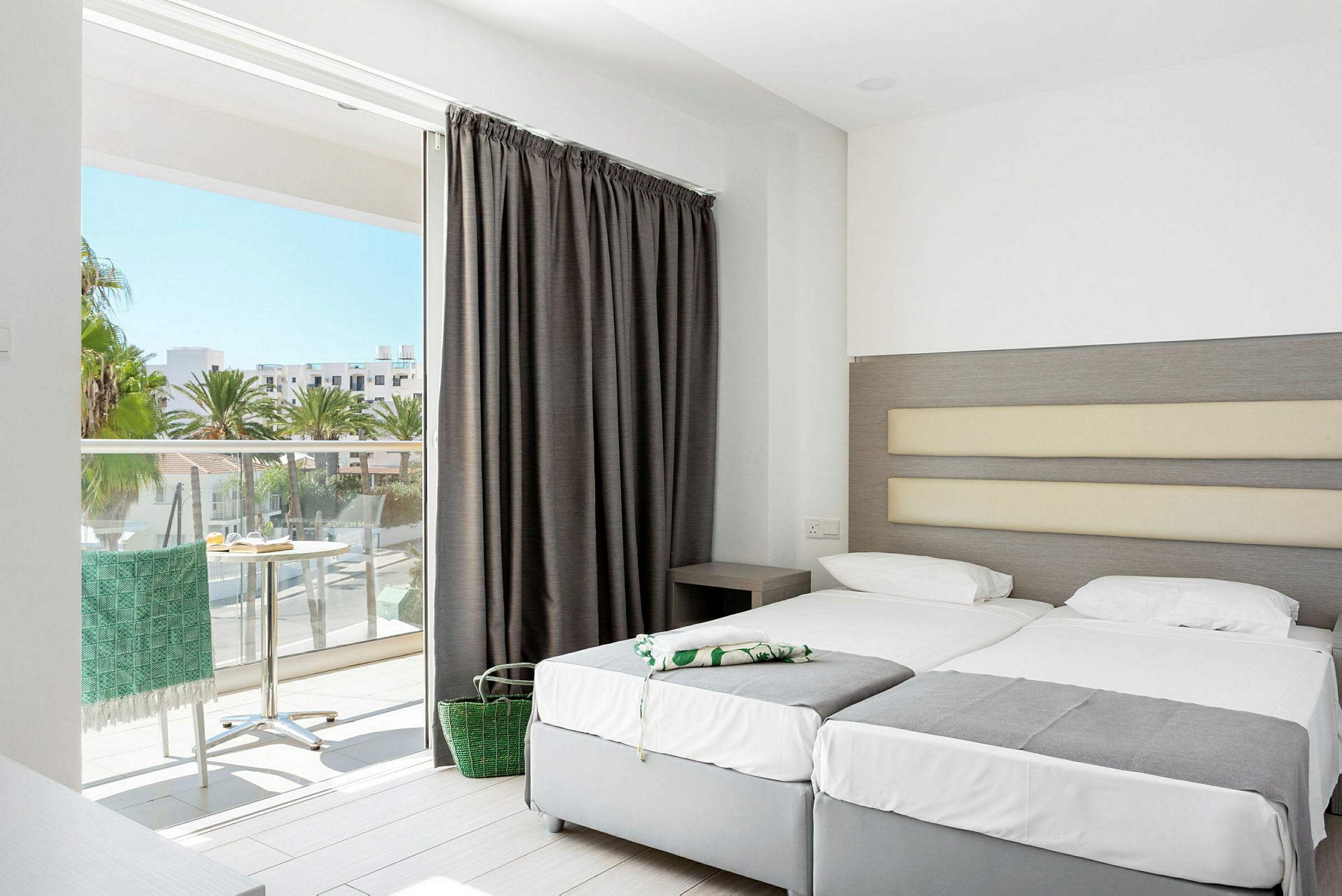 Hotellrum med två bäddade sängar och utsikt från ett öppet fönster mot palmer och hus. (AI-genererad text)