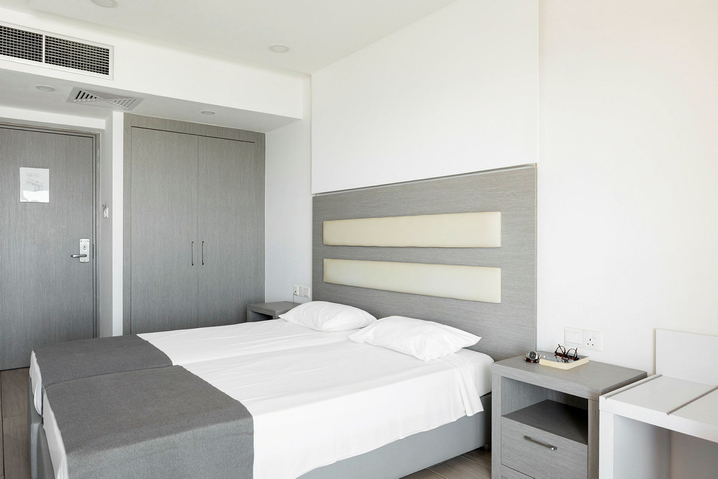 Bild visar ett hotellrum med två enkelsängar, grå inredning och vita väggar. (AI-genererad text)