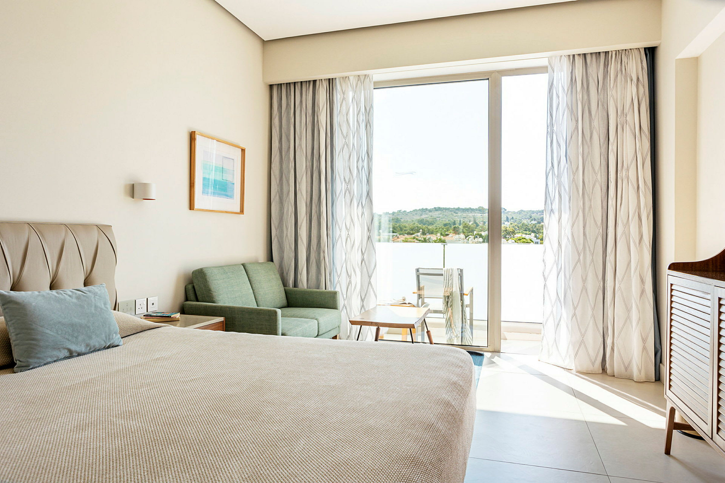 Ett hotellrum med en säng, soffa och bord, samt en balkong med stolar och utsikt över grönska. (AI-genererad text)