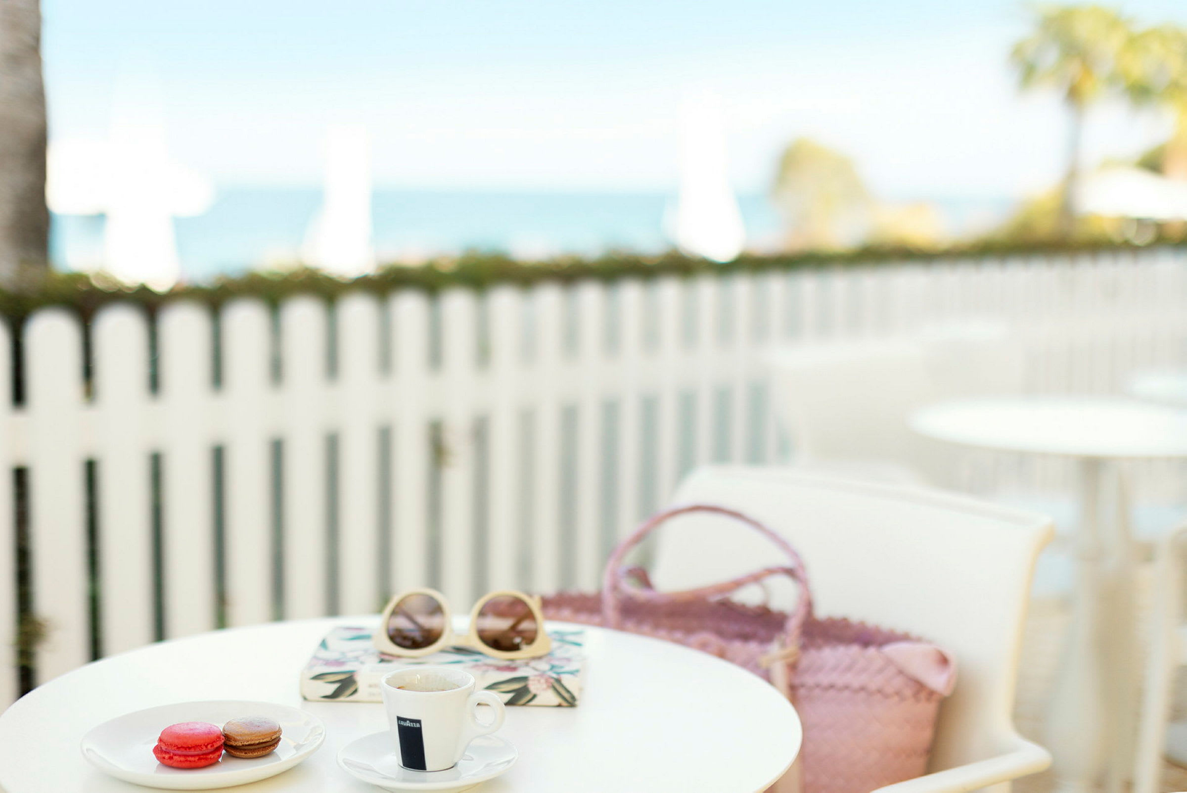 En kopp kaffe, macarons, solglasögon och en väska på ett cafébord med en vit trästaket och havet i bakgrunden. (AI-genererad text)