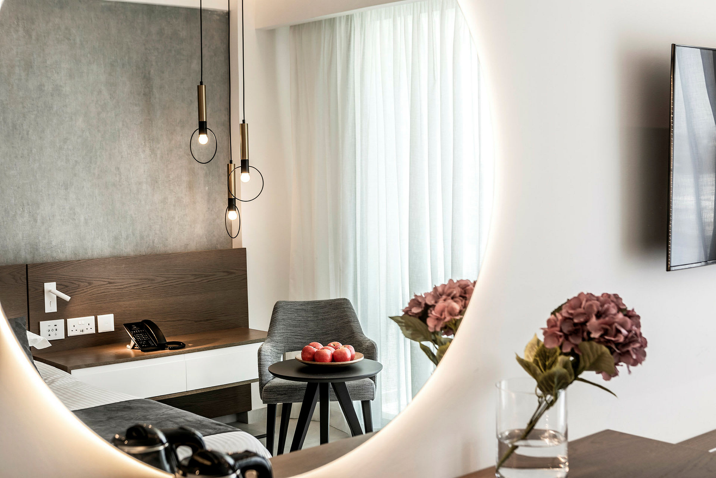 Hotellrumsinteriör med en spegel som reflekterar en säng, ett nattduksbord med en telefon, en skål med röda äpplen på ett runt bord samt en vas med lila blommor. (AI-genererad text)