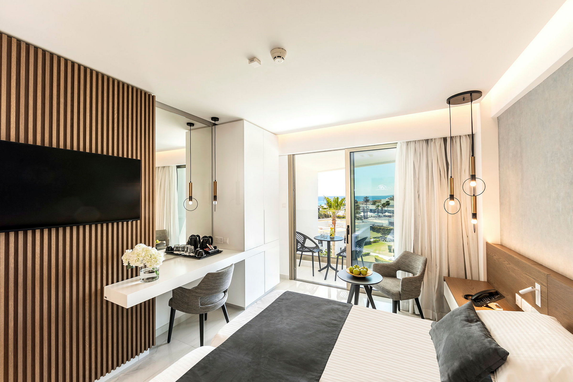 Modern hotellrum med en dubbelsäng, bord och stolar samt utsikt över havet från en balkong. (AI-genererad text)