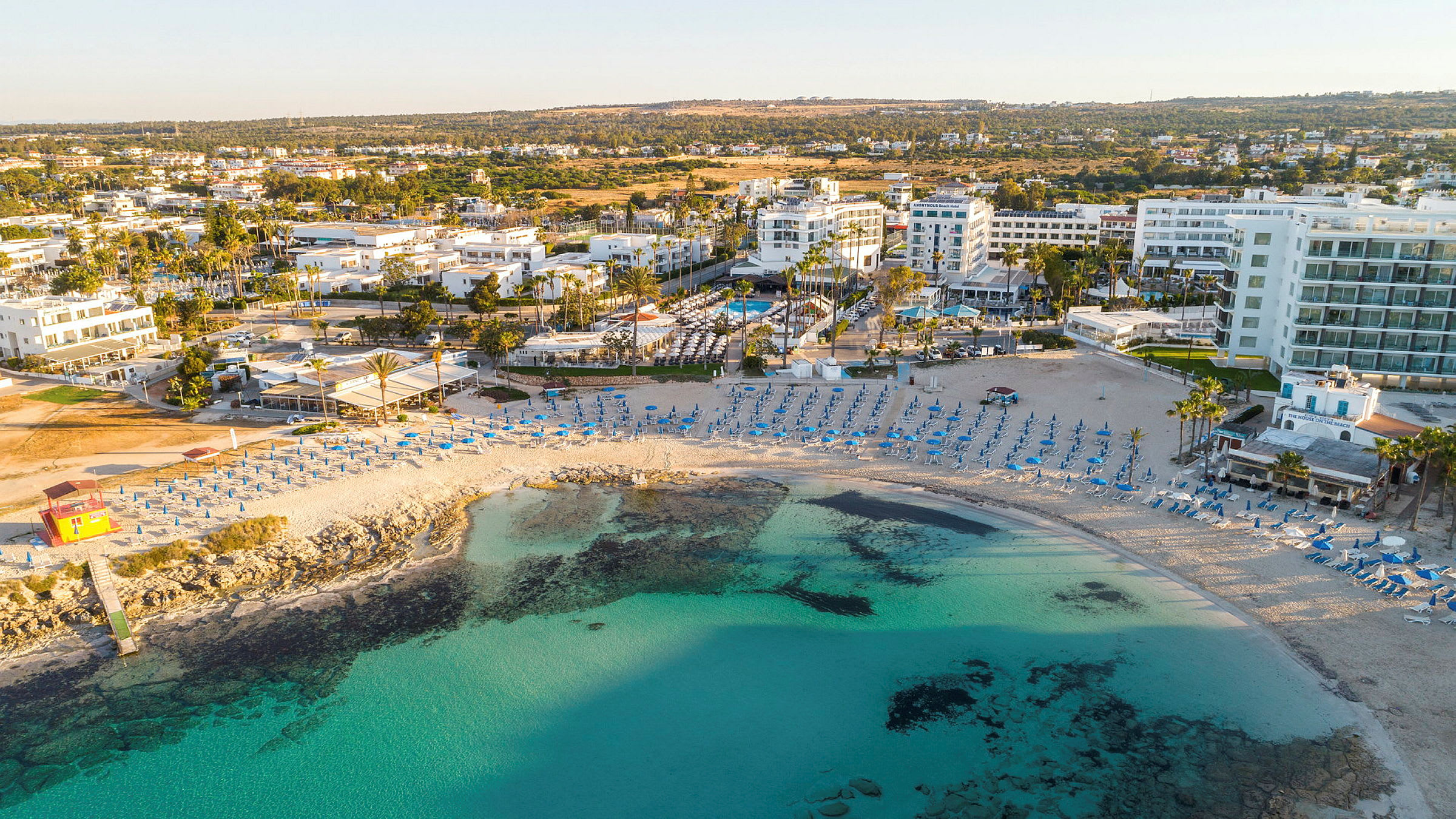 Strand med parasoller och stolar, omgiven av turkost vatten, byggnader och grönska. (AI-genererad text)