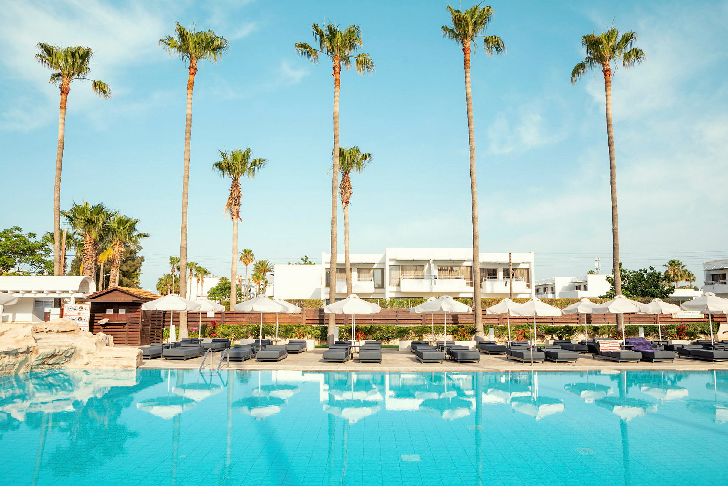 Pool med solstolar och parasoller, omgiven av palmer och vita byggnader. (AI-genererad text)