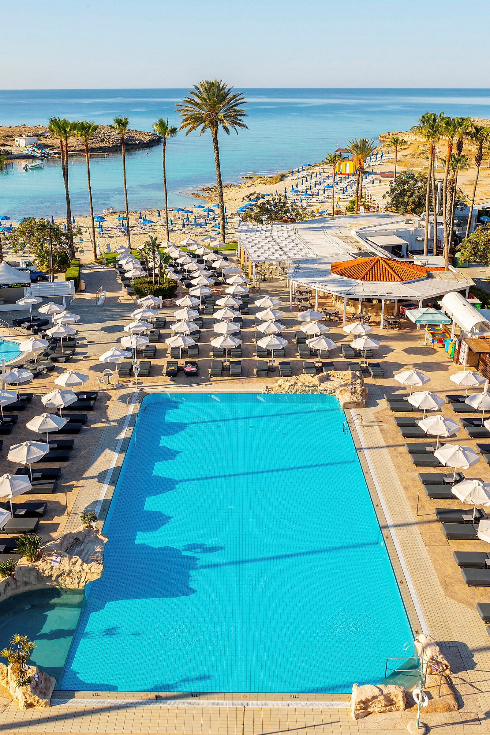 Bild som visar en swimmingpool med solstolar och parasoller runt omkring, samt en strand och hav i bakgrunden. (AI-genererad text)