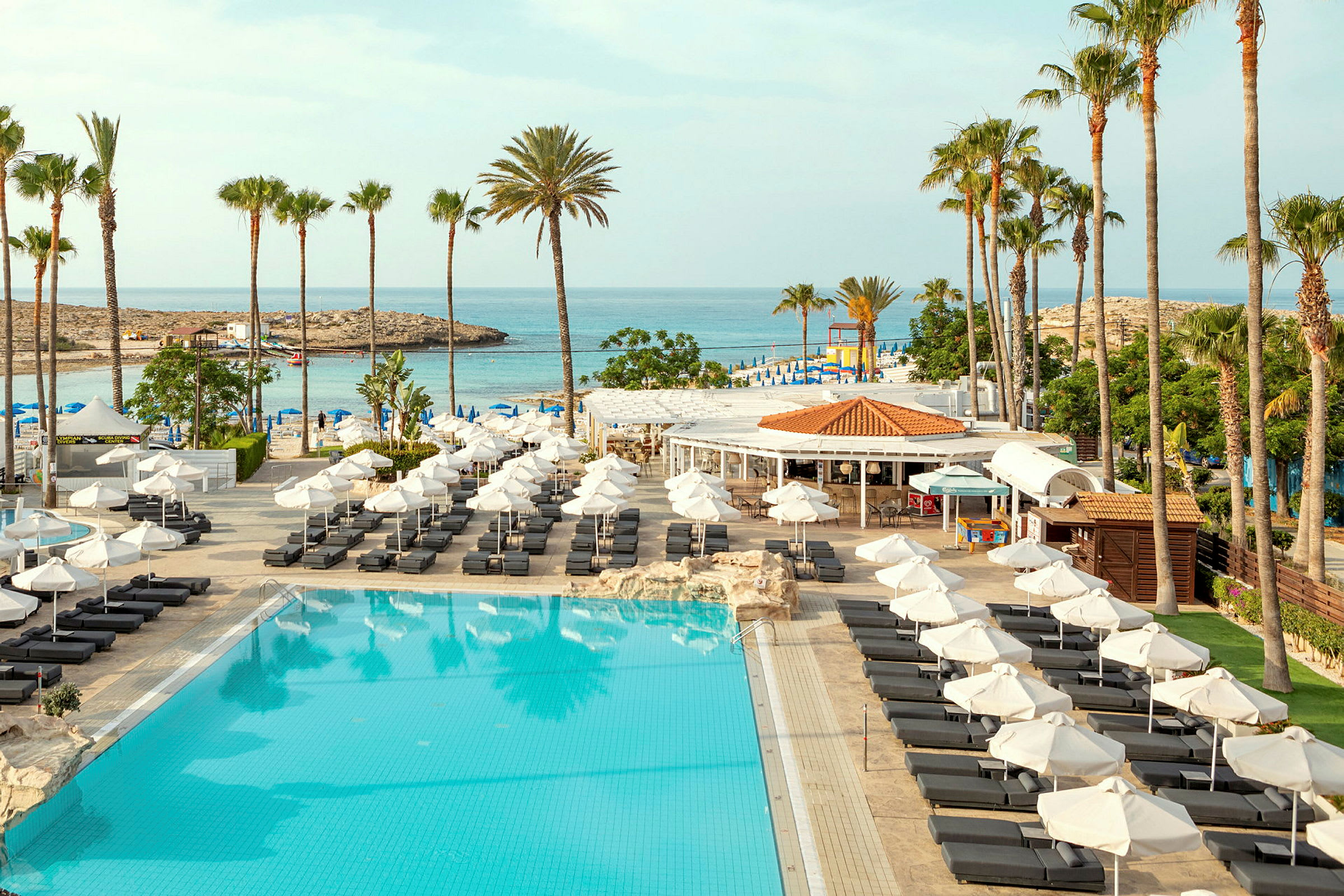 Bild av en resort med en stor pool, solstolar och palmer. I bakgrunden syns havet och en strand. (AI-genererad text)