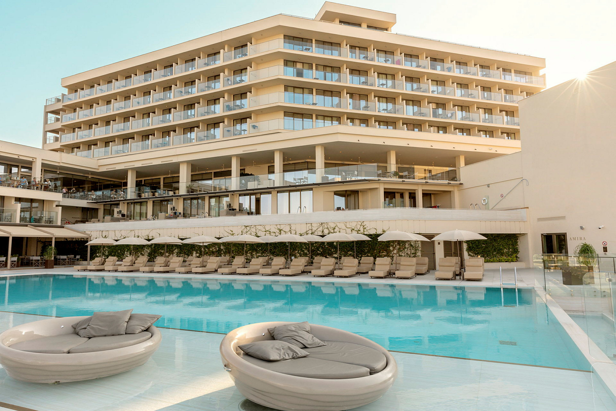 Ett hotell med flera våningar och stora balkonger syns bakom en swimmingpool med solstolar och parasoller. (AI-genererad text)