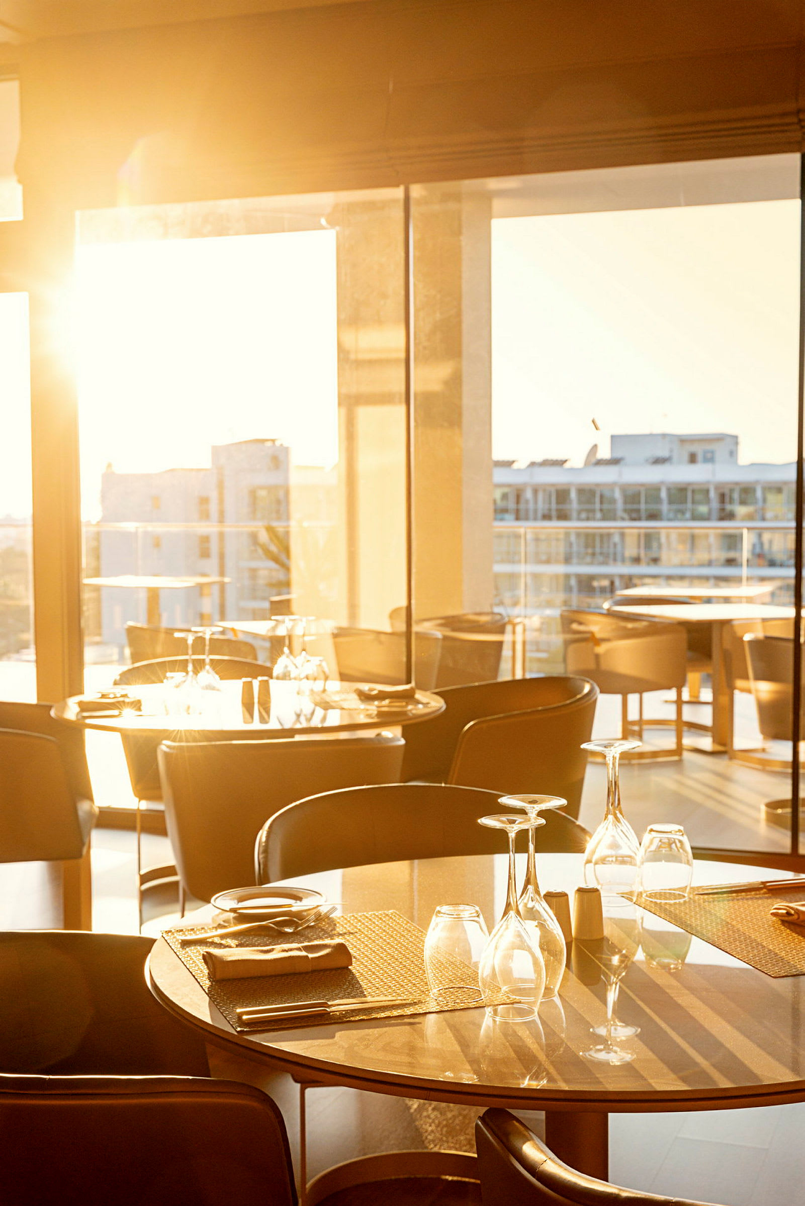Ett dukat bord i ett soligt rum med stora fönster och utsikt över andra byggnader. (AI-genererad text)