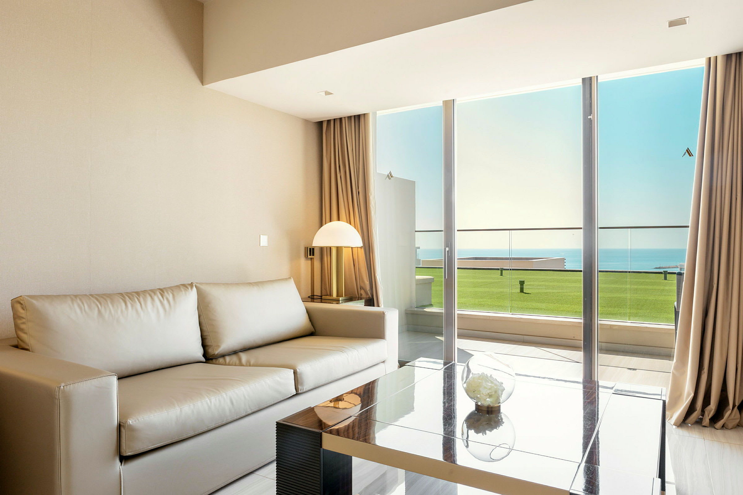 Bild på ett modernt vardagsrum med en beige soffa, glasbord och en stor fönsterdörr som vetter mot en balkong med utsikt mot havet. (AI-genererad text)