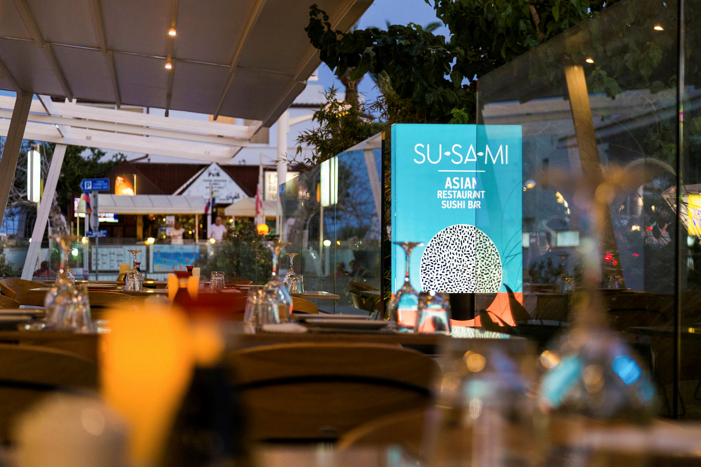 Uteservering med dukade bord och en skylt med texten 'Su-Sa-Mi Asian Restaurant Sushi Bar'. (AI-genererad text)