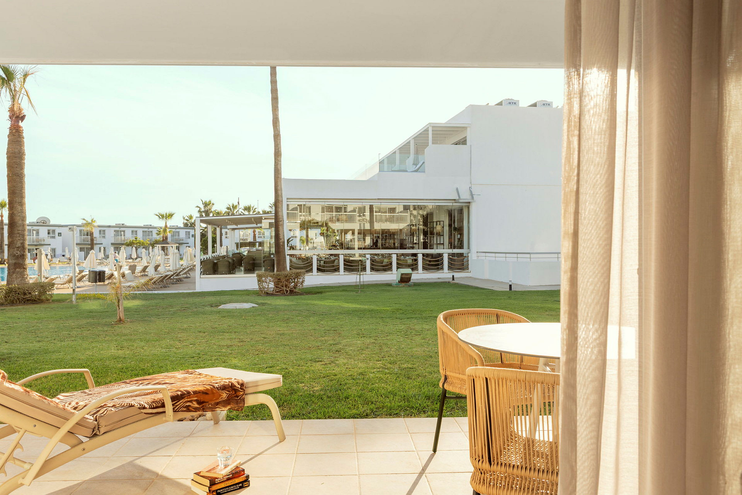 Utsikt från en terrass med en solstol och bord, med en grön gräsmatta och hotellbyggnader i bakgrunden. (AI-genererad text)