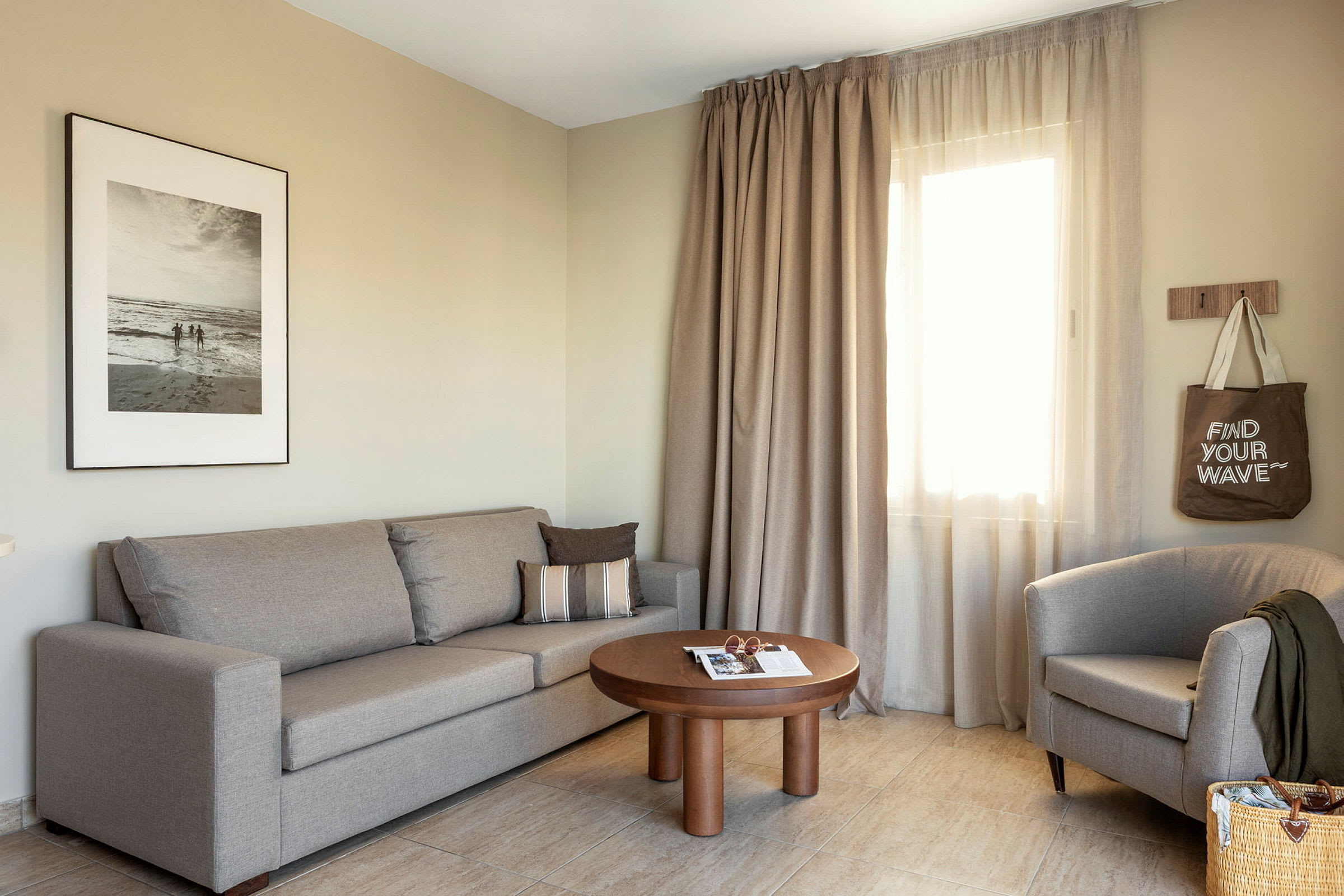 Ett vardagsrum med en beige soffa, en fåtölj, ett träbord och gardiner vid ett fönster. (AI-genererad text)