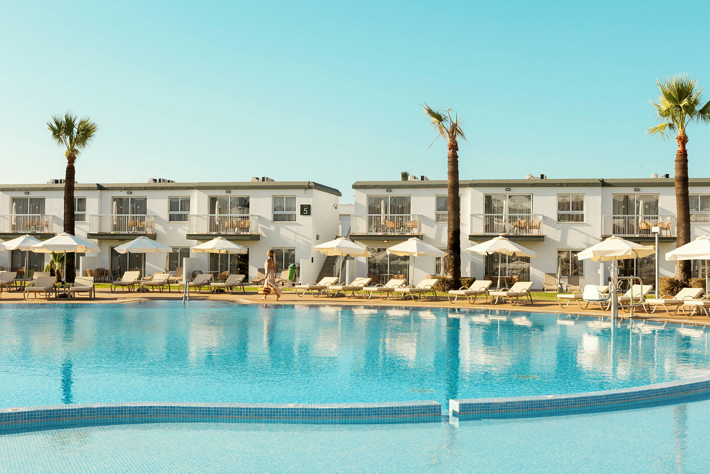 Hotellbyggnad med balkonger och en pool framför, omgiven av solsängar och parasoller. (AI-genererad text)