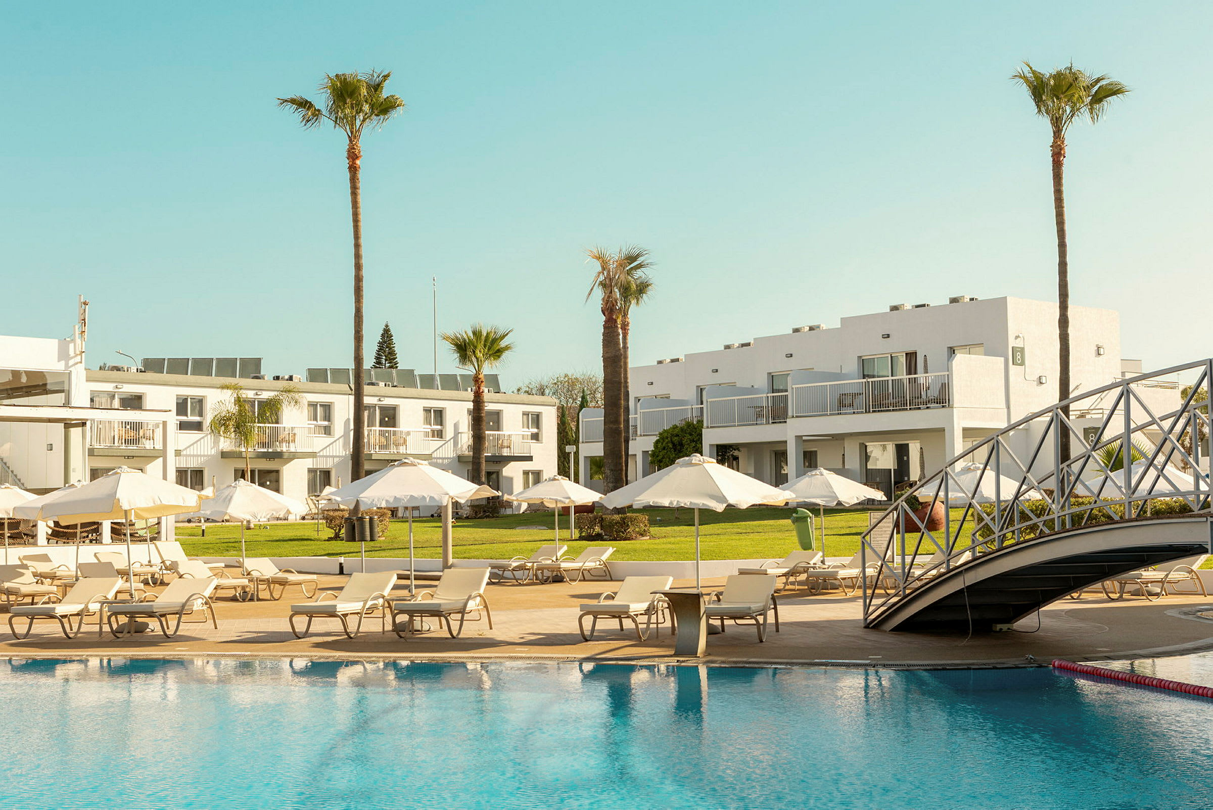 Hotellområde med pool, solstolar och parasoller samt vita byggnader i bakgrunden. (AI-genererad text)