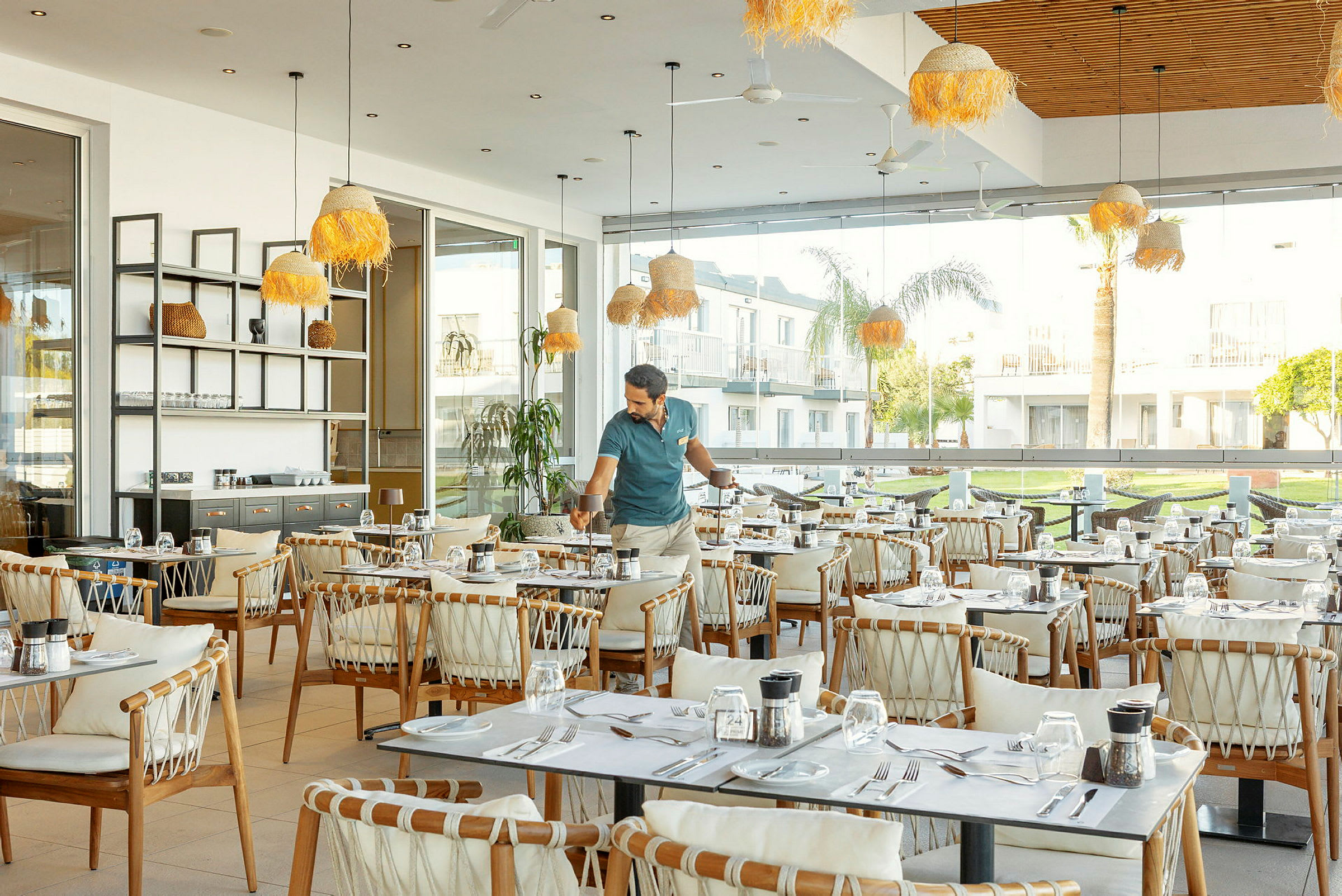 Restaurang med bord och stolar samt stora fönster som vetter mot en grön utomhusmiljö. (AI-genererad text)