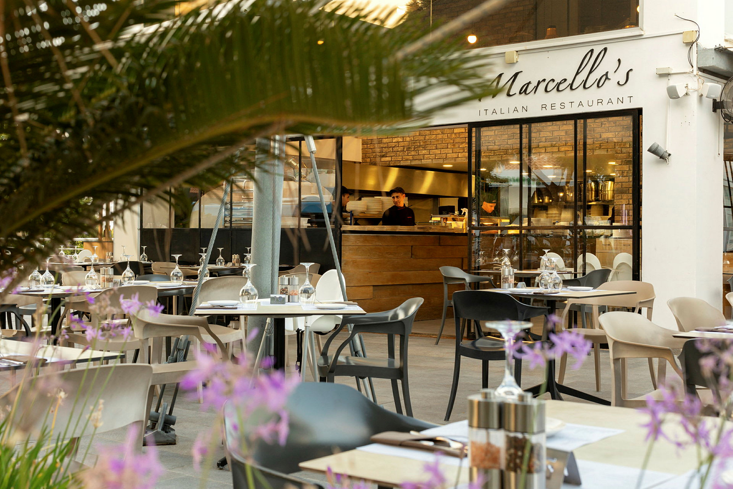 Uteservering med bord och stolar framför en italiensk restaurang med glasväggar och ett träserveringsområde. (AI-genererad text)