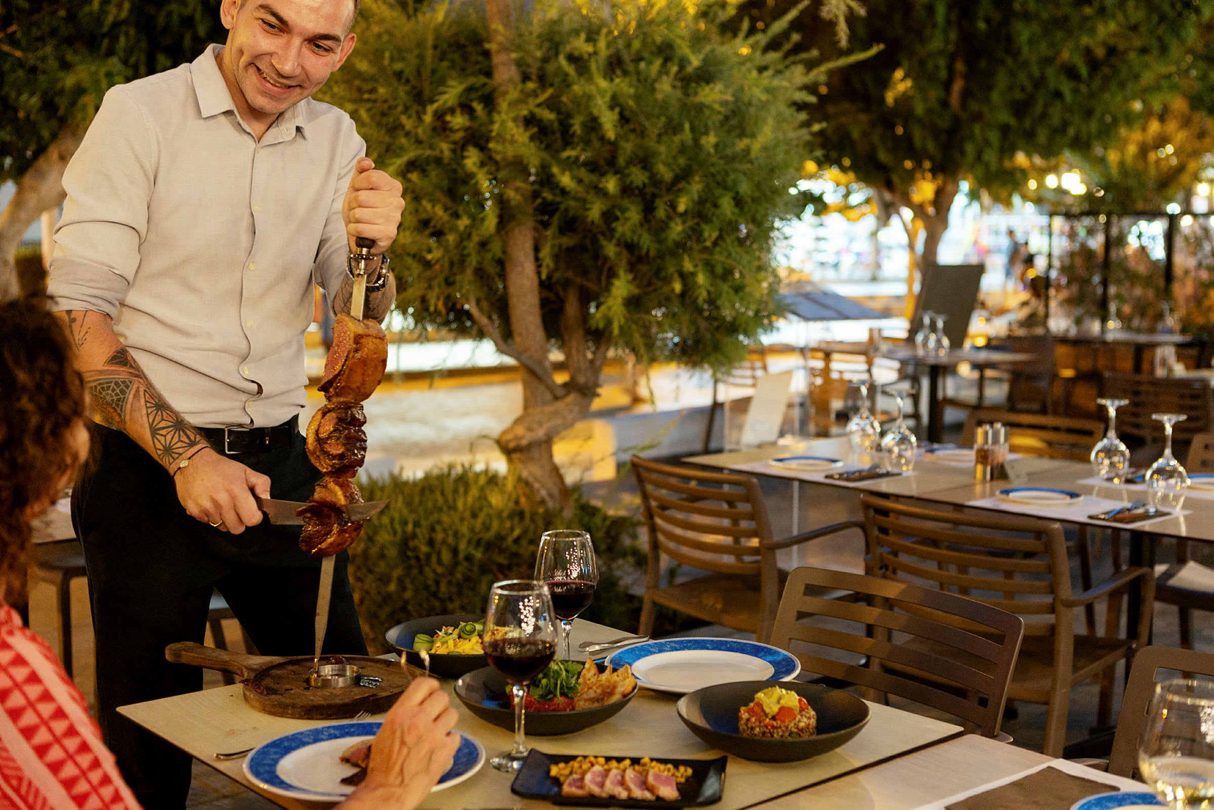 En person serverar grillat kött vid ett bord utomhus. Rätter och glas finns på bordet. (AI-genererad text)