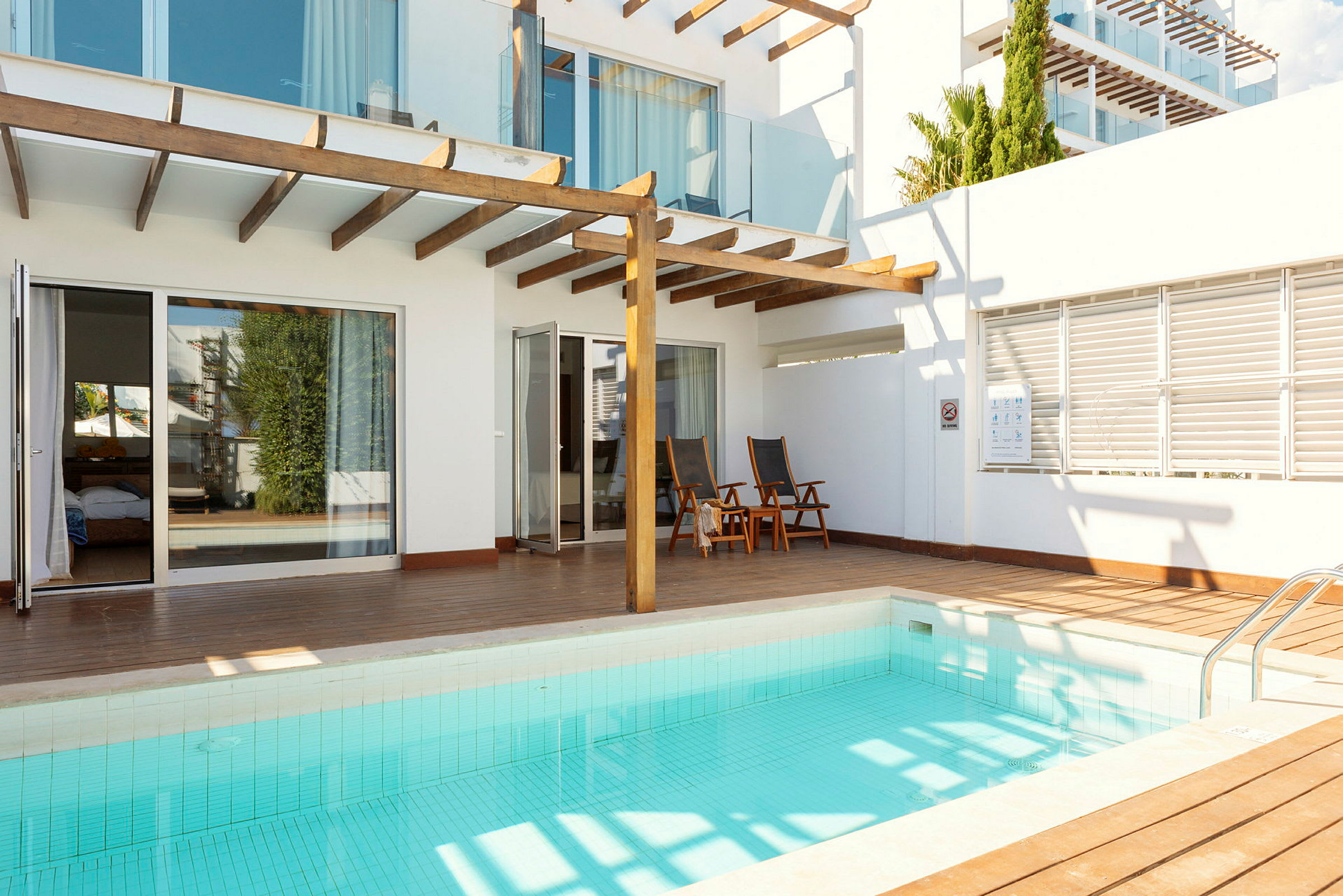 En mindre swimmingpool med en trädterrass och moderna vita byggnader i bakgrunden. (AI-genererad text)