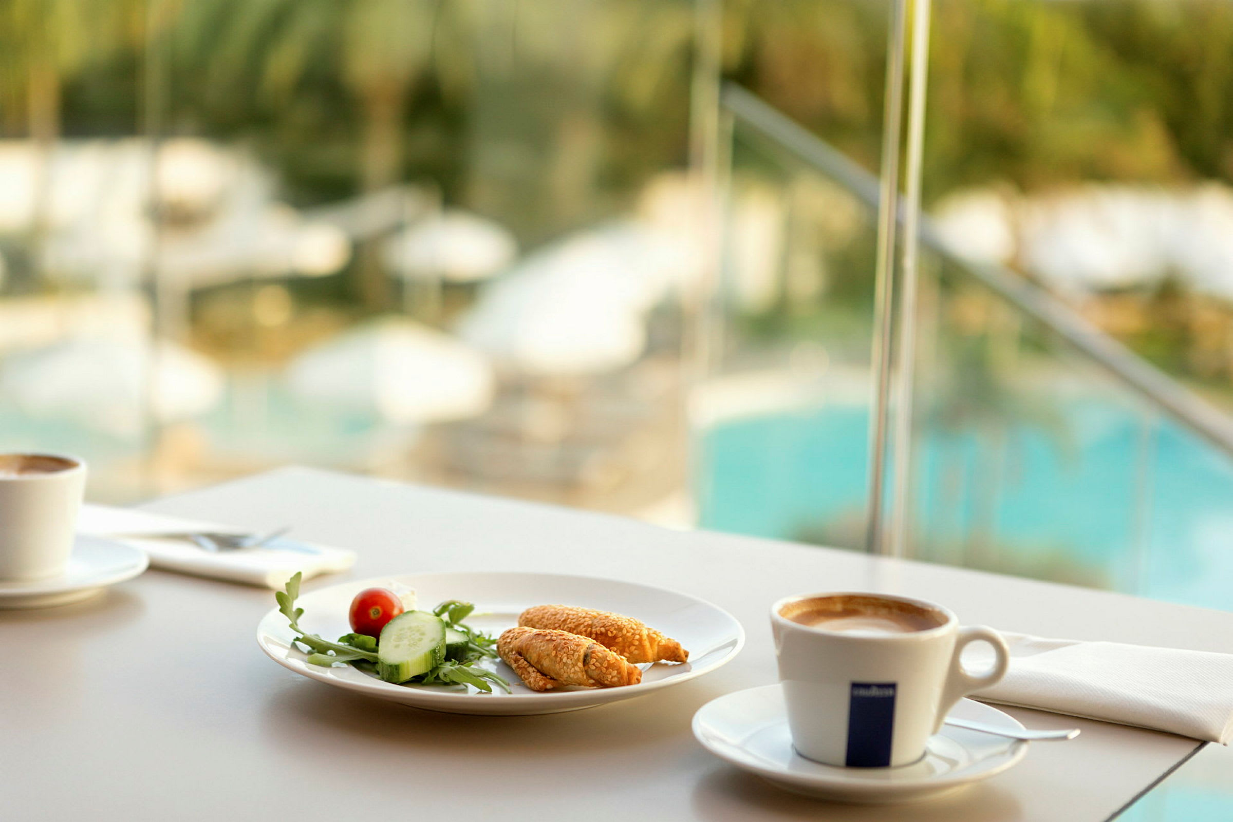 Frukostservering med kaffe och ett fat med grönsaker och bakverk, bakgrund med utsikt över en pool. (AI-genererad text)