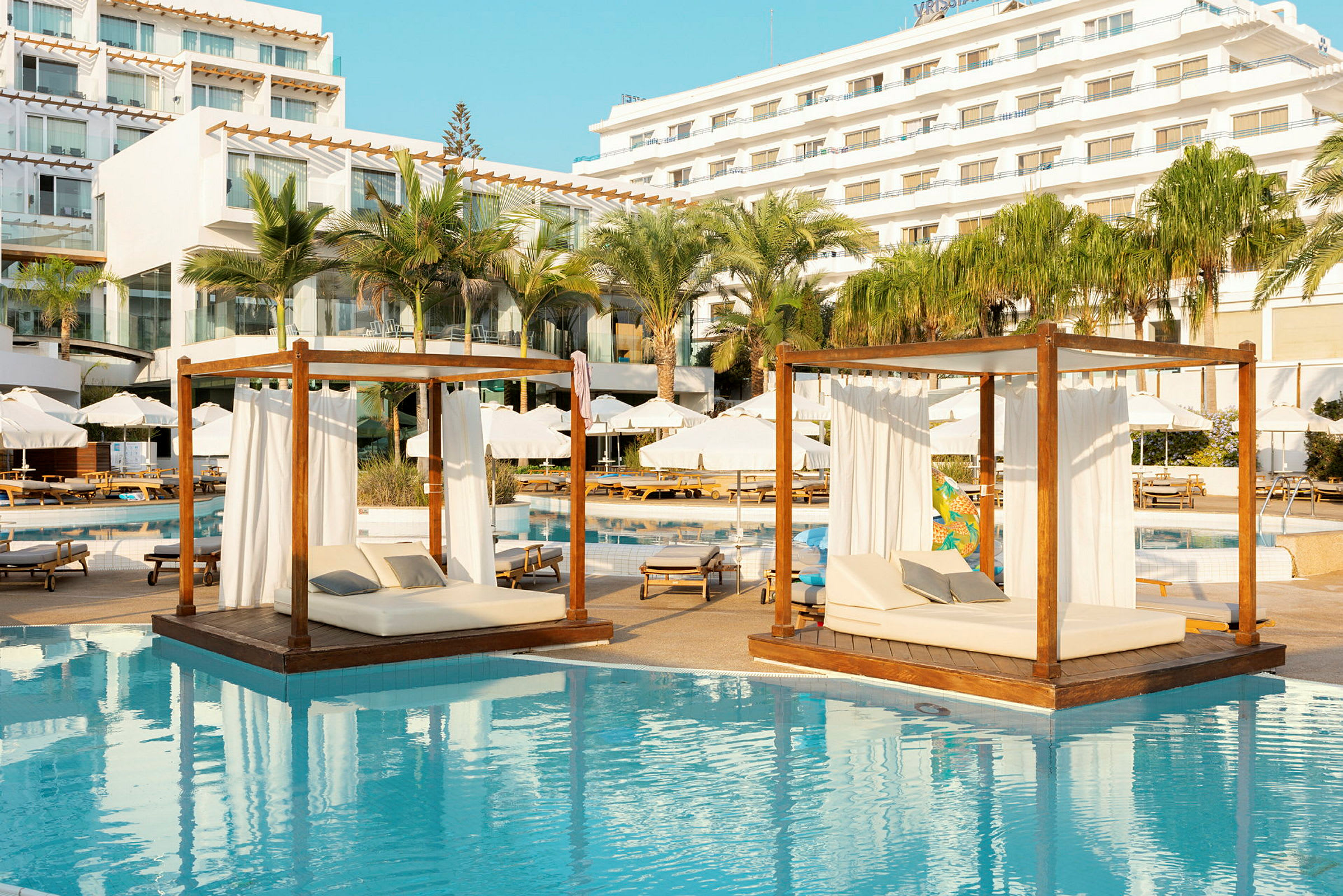 Liggande dagbäddar med vita draperier över vatten vid en solig hotellpool. (AI-genererad text)