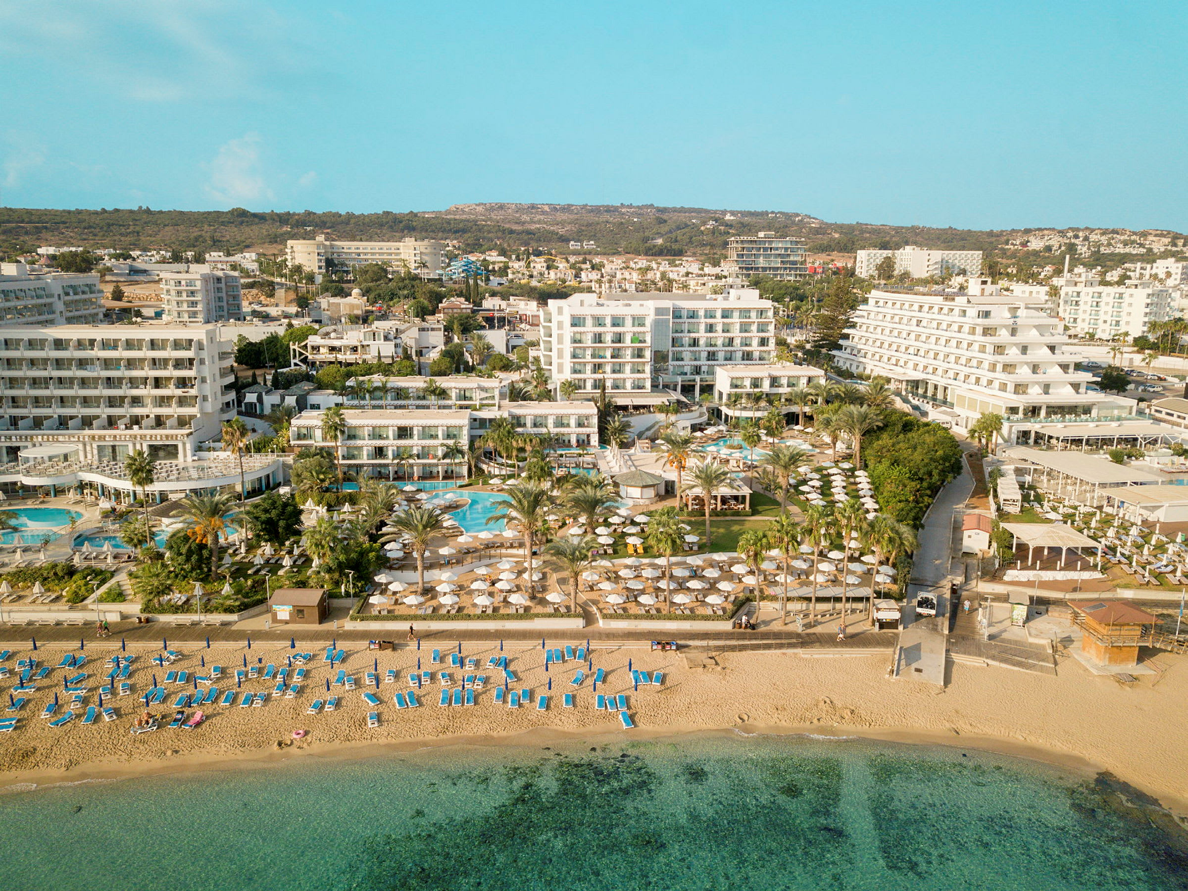 Flygfoto som visar en strand med solstolar och parasoller, samt hotellbyggnader och poolområde i bakgrunden. (AI-genererad text)