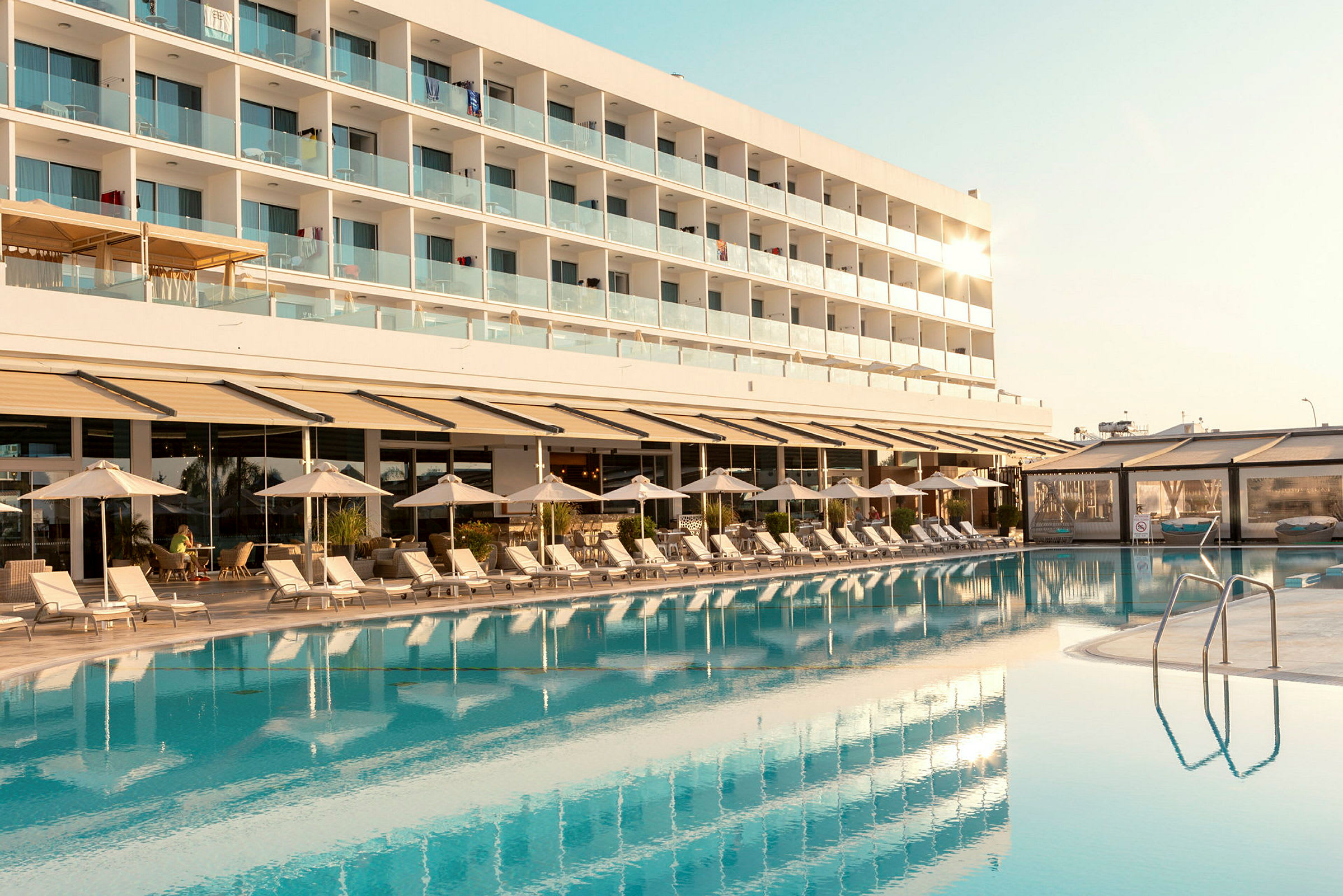 En hotellbyggnad med balkonger syns bakom en pool och solstolar. (AI-genererad text)