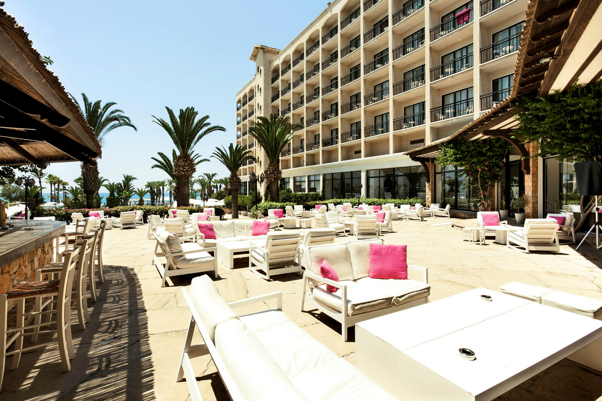 En rymlig solterrass med utemöbler i vitt och rosa, omgiven av palmer och ett stort hotell i bakgrunden. (AI-genererad text)