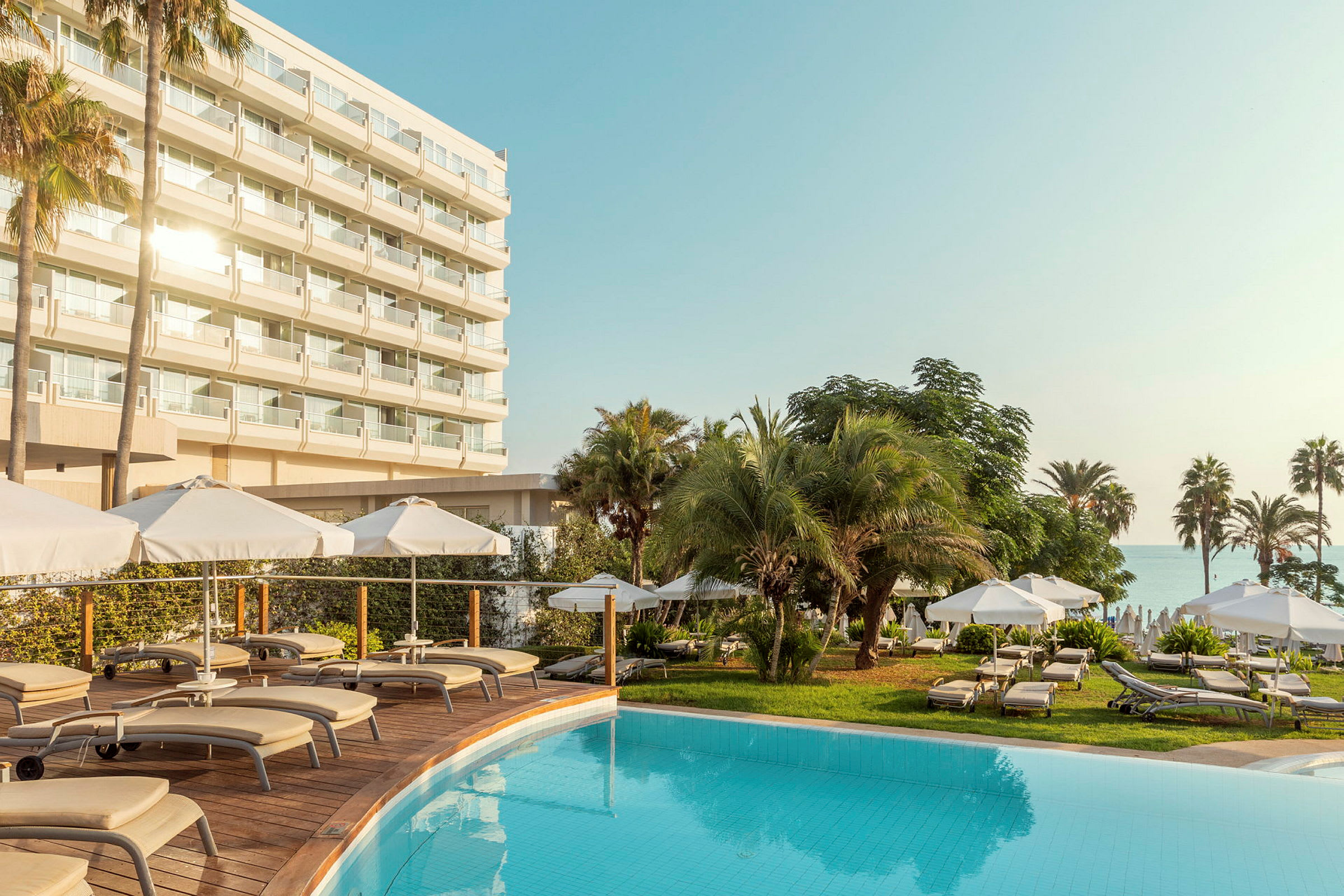 Ett hotell med flera våningar bakom en pool och solstolar omgivna av parasoller och palmer. (AI-genererad text)