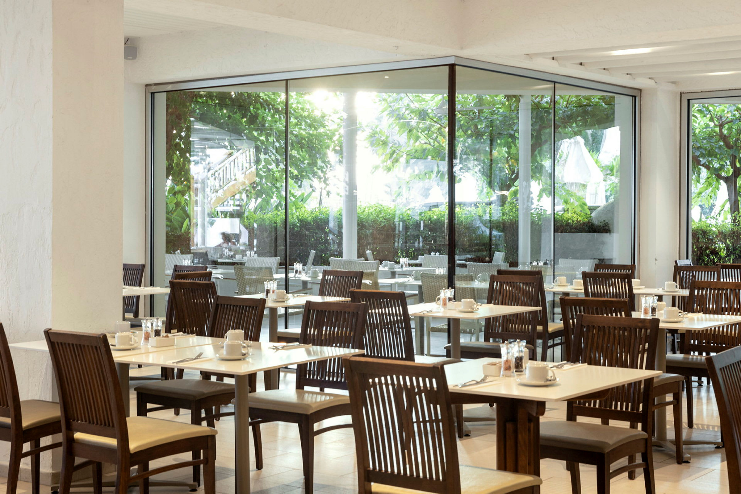 Bild av en restaurang med träbord och stolar, samt utsikt mot grönska genom stora glasfönster. (AI-genererad text)