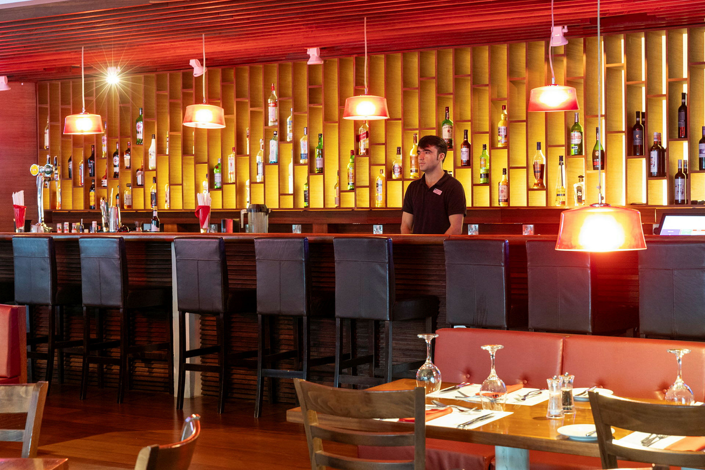 En modern bar med stolar, flaskor på väggen och matbord framför. (AI-genererad text)