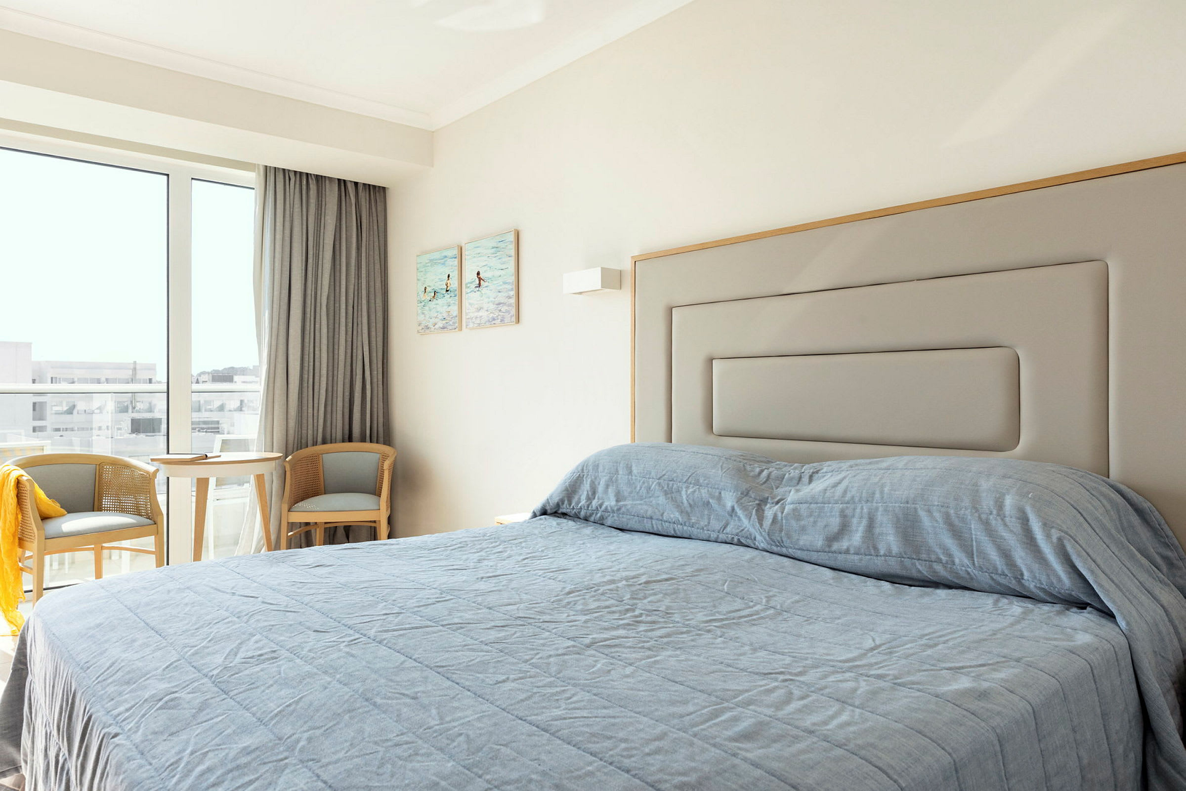 Ett hotellrum med en säng bäddad med blått överkast, två stolar och ett bord nära ett fönster. (AI-genererad text)