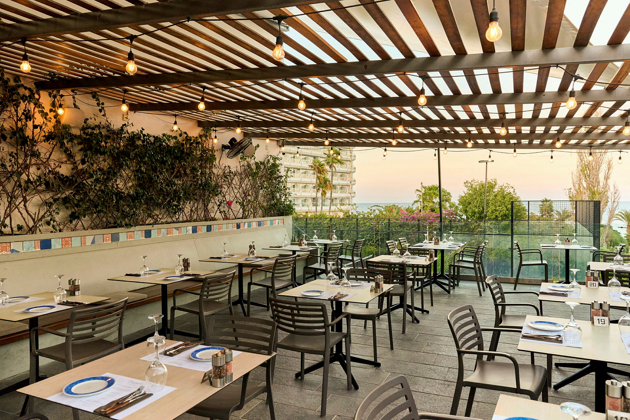 En utomhusrestaurang med dukade bord och stolar under en träpergola dekorerad med växter och ljusslingor. Utsikt över en byggnad och trädgård mot havet. (AI-genererad text)