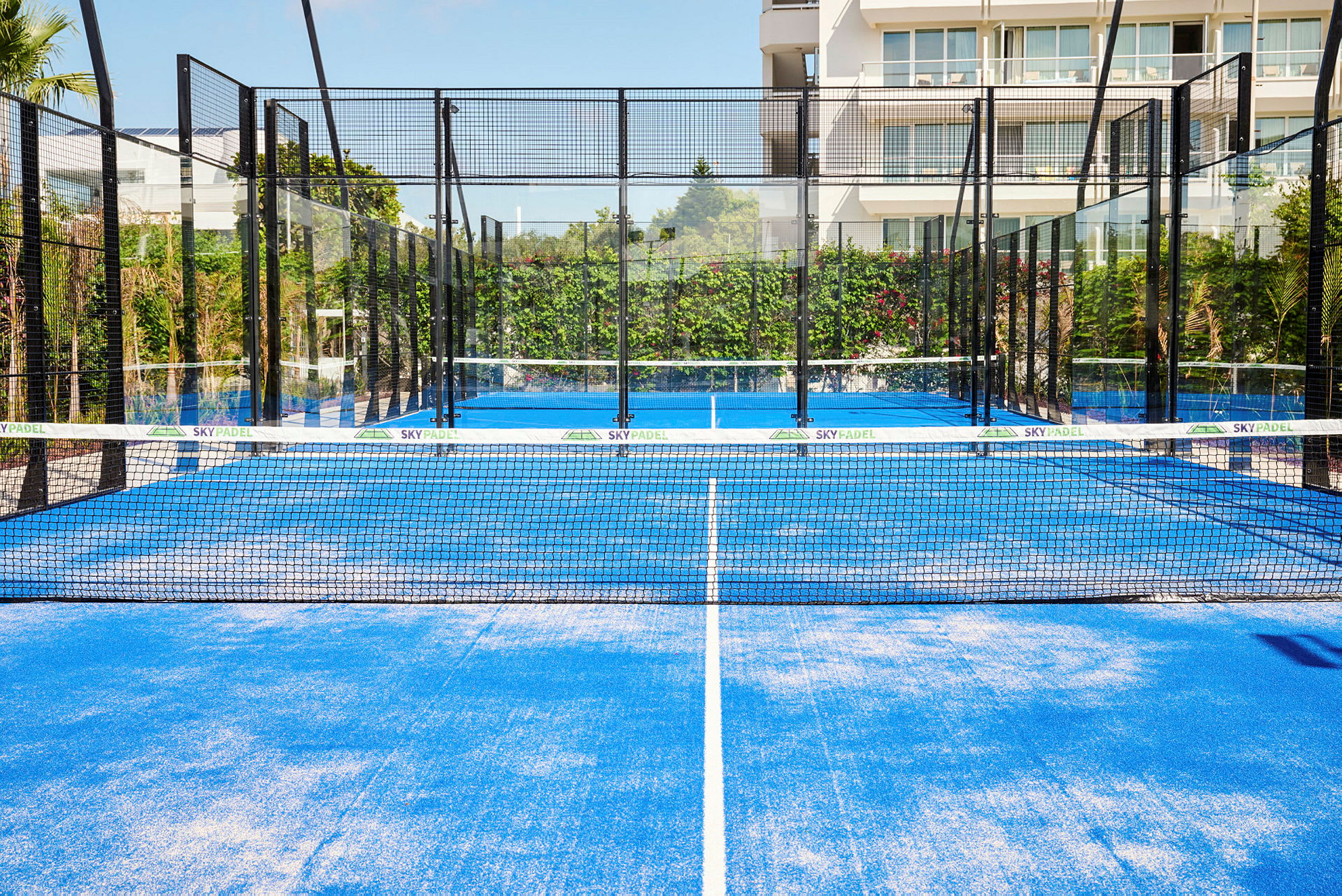 En blå padelbana med nät och omgivande glasväggar, belägen utomhus med grönska och en byggnad i bakgrunden. (AI-genererad text)