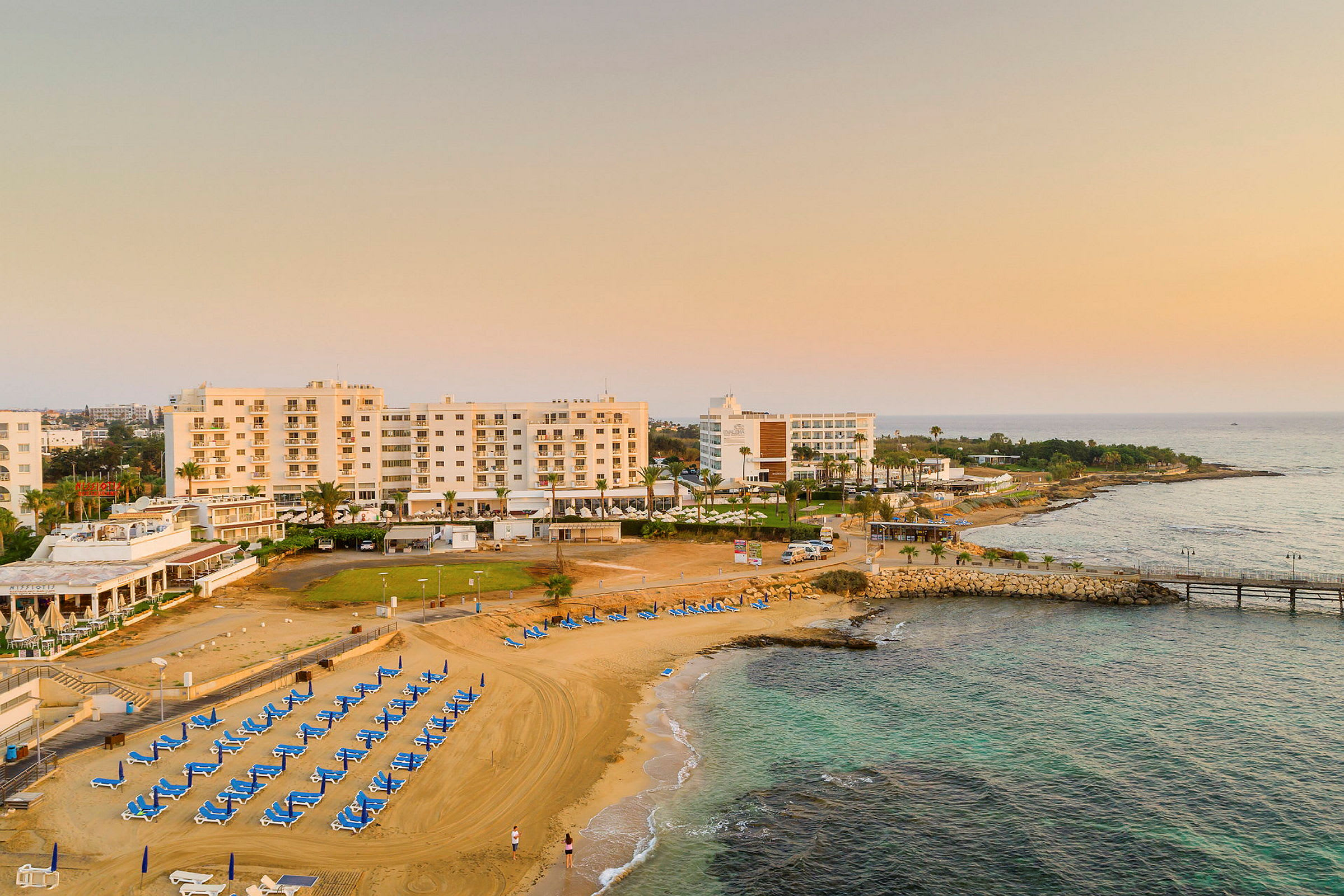 Flygfoto över hotellbyggnader vid en strand med solstolar och havet i bakgrunden. (AI-genererad text)