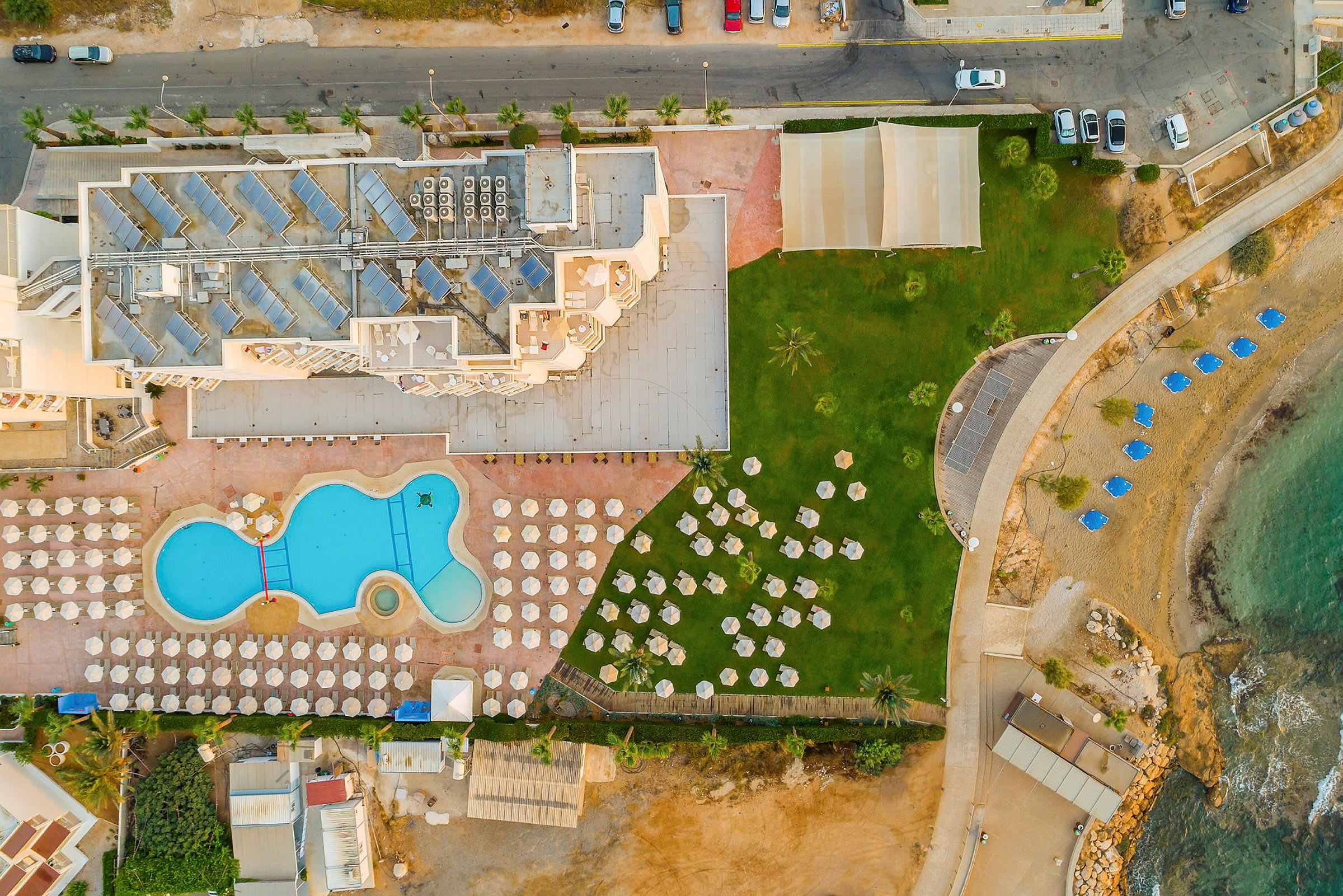 Flygfoto av hotellområde med pool, solstolar, gräsområde och strand med parasoller. (AI-genererad text)