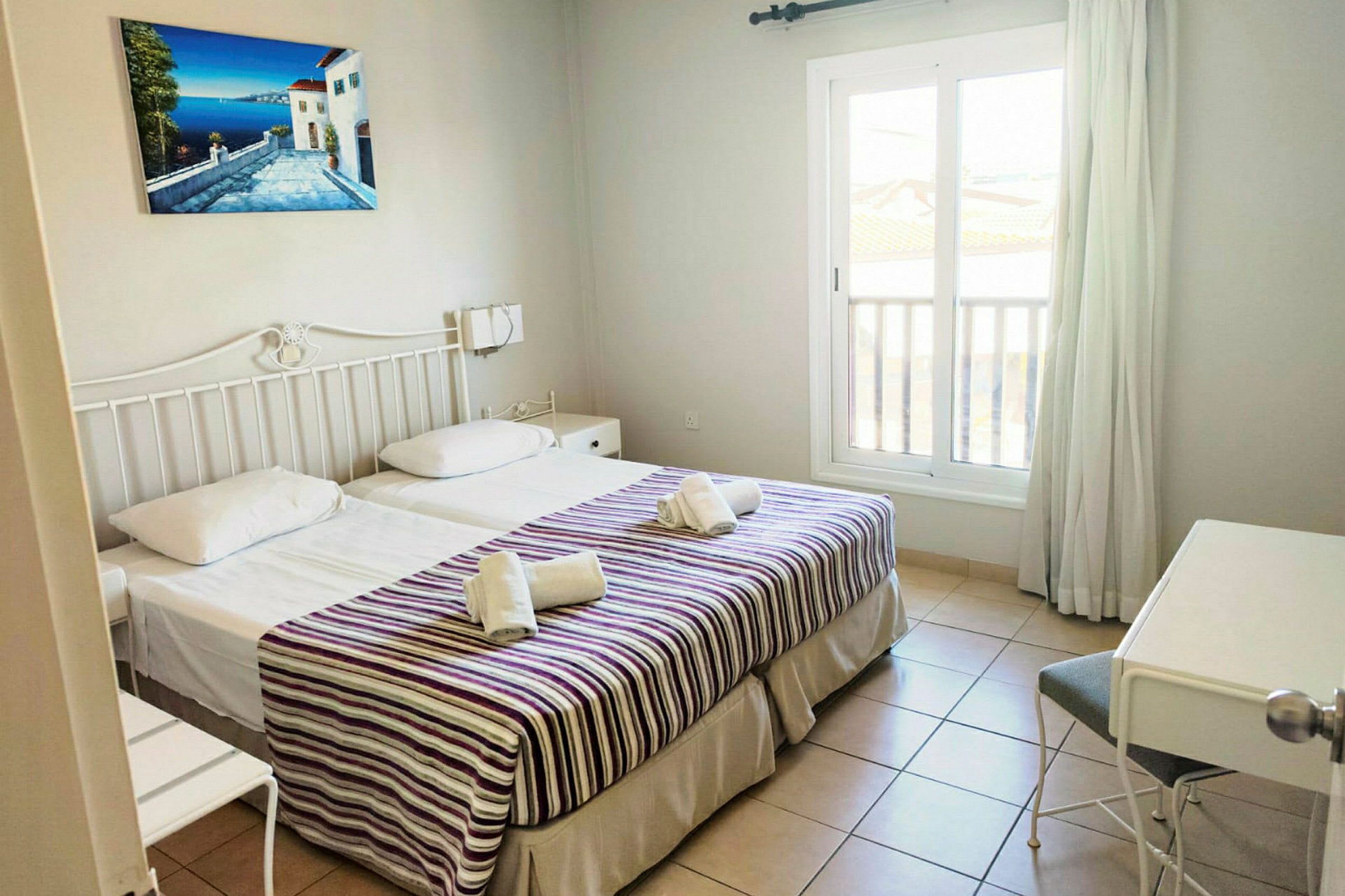 Ett hotellrum med två enkelsängar, dekorerade med randiga överkast och handdukar. Ett fönster och en skrivbordshörna är synliga. (AI-genererad text)