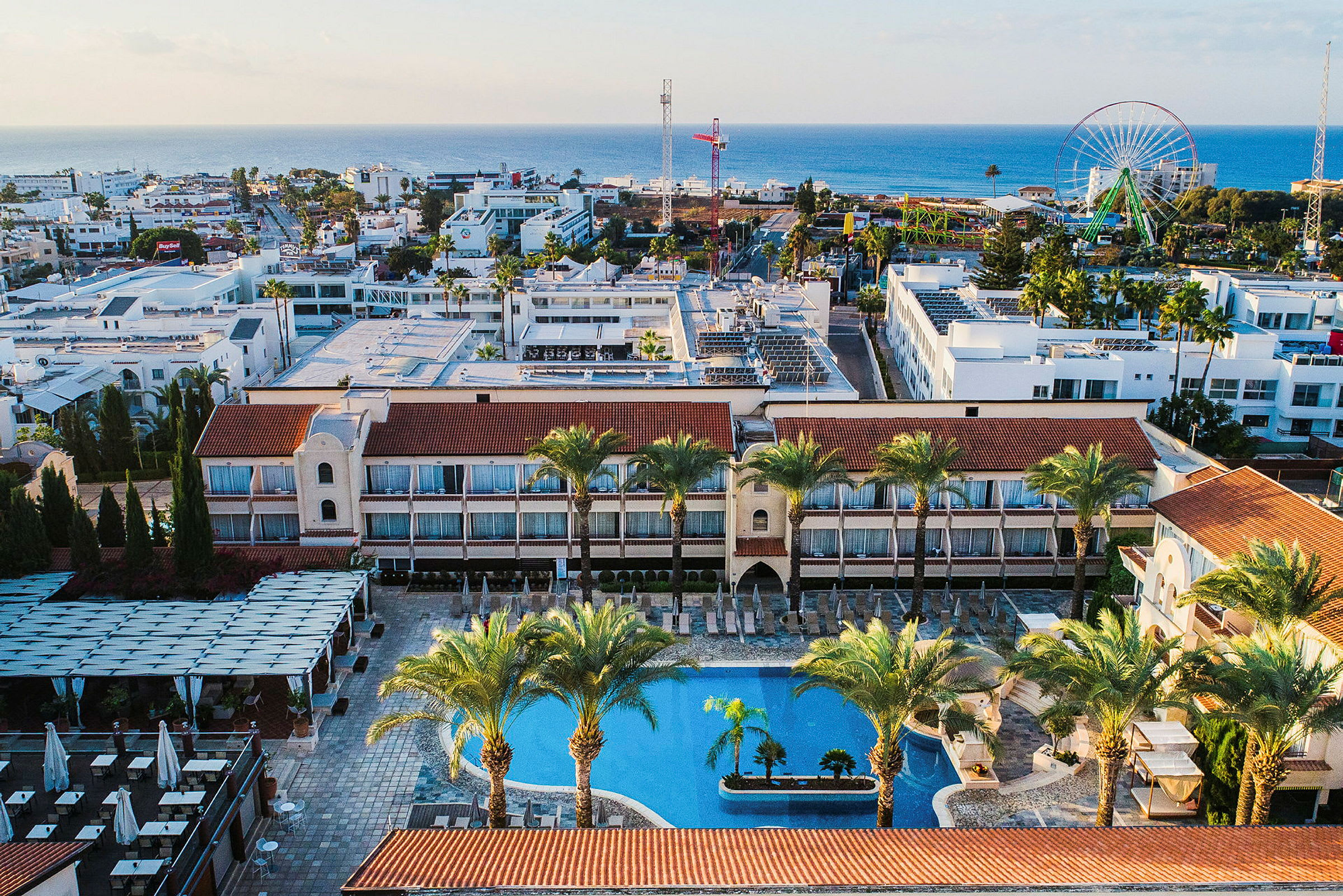Flygfoto av hotellkomplex med pool, palmer och omgivande byggnader nära stranden. (AI-genererad text)