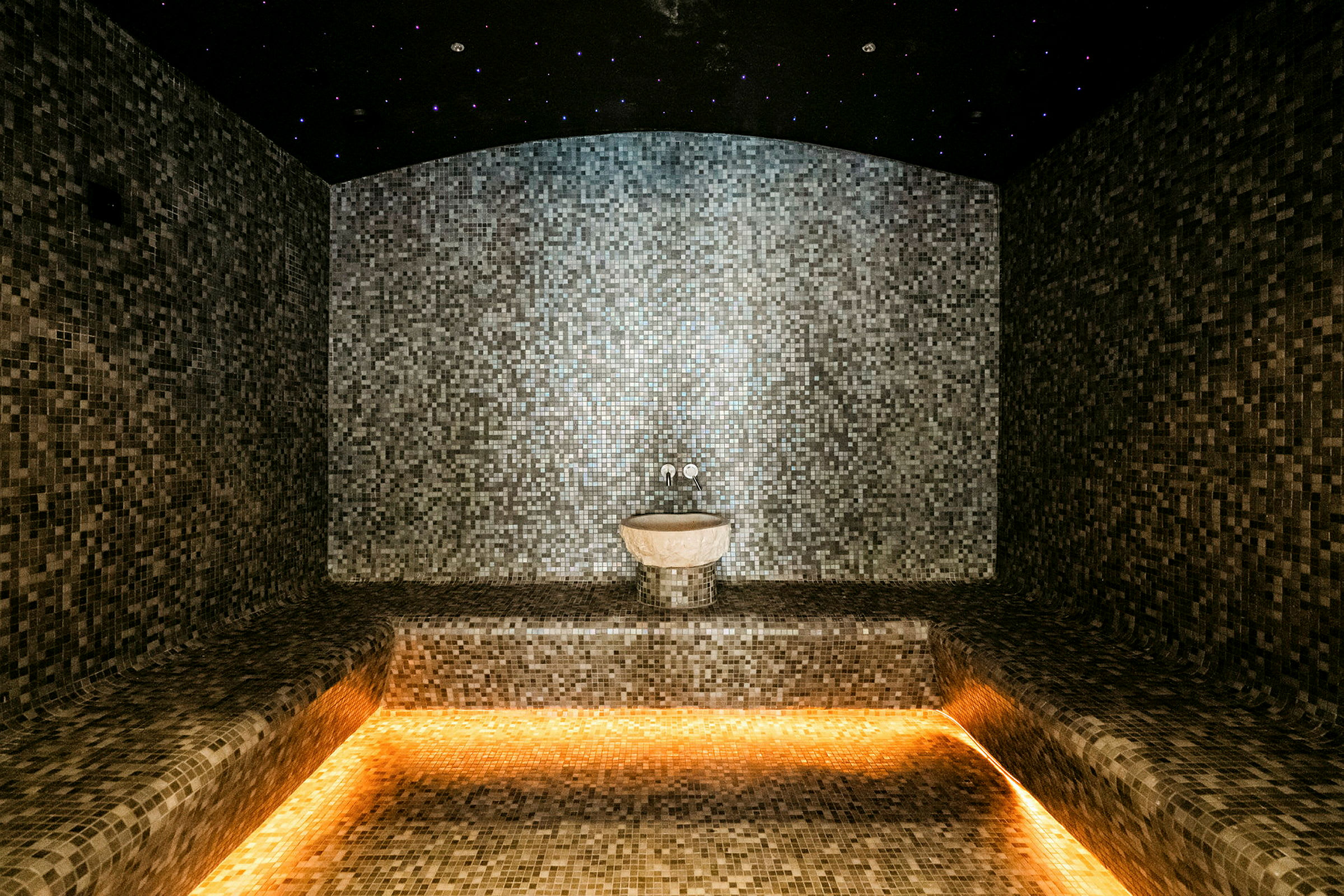 Ett spa-rum med mosaikväggar och en stjärnbelyst takdesign, med en liten tvättskål vid väggen. (AI-genererad text)