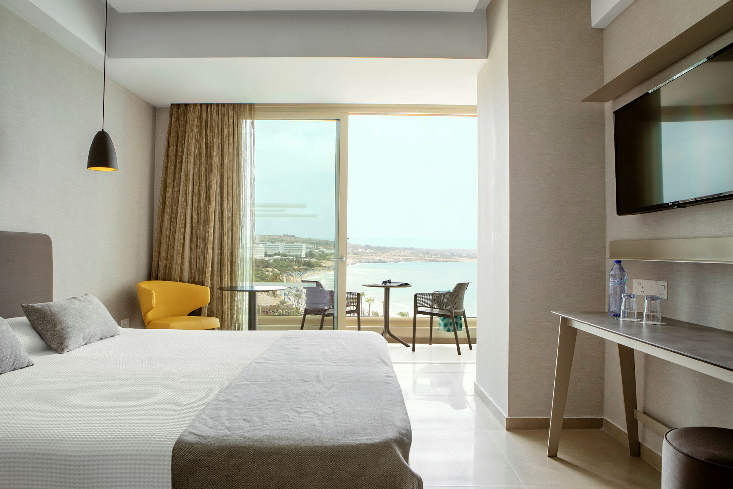 Ett hotellrum med modern inredning, en bred säng, en gul sittstol, ett bord med utsikt över stranden genom stora fönster. (AI-genererad text)