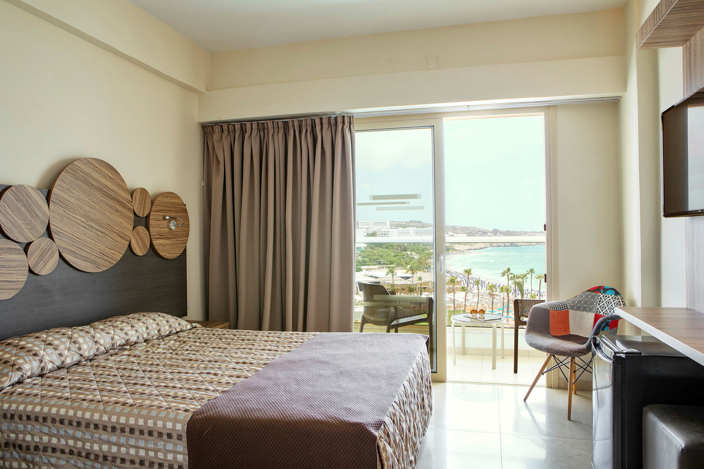 Ett hotellrum med en dubbelsäng, skrivbord och stolar, med utsikt över stranden från balkongdörren. (AI-genererad text)