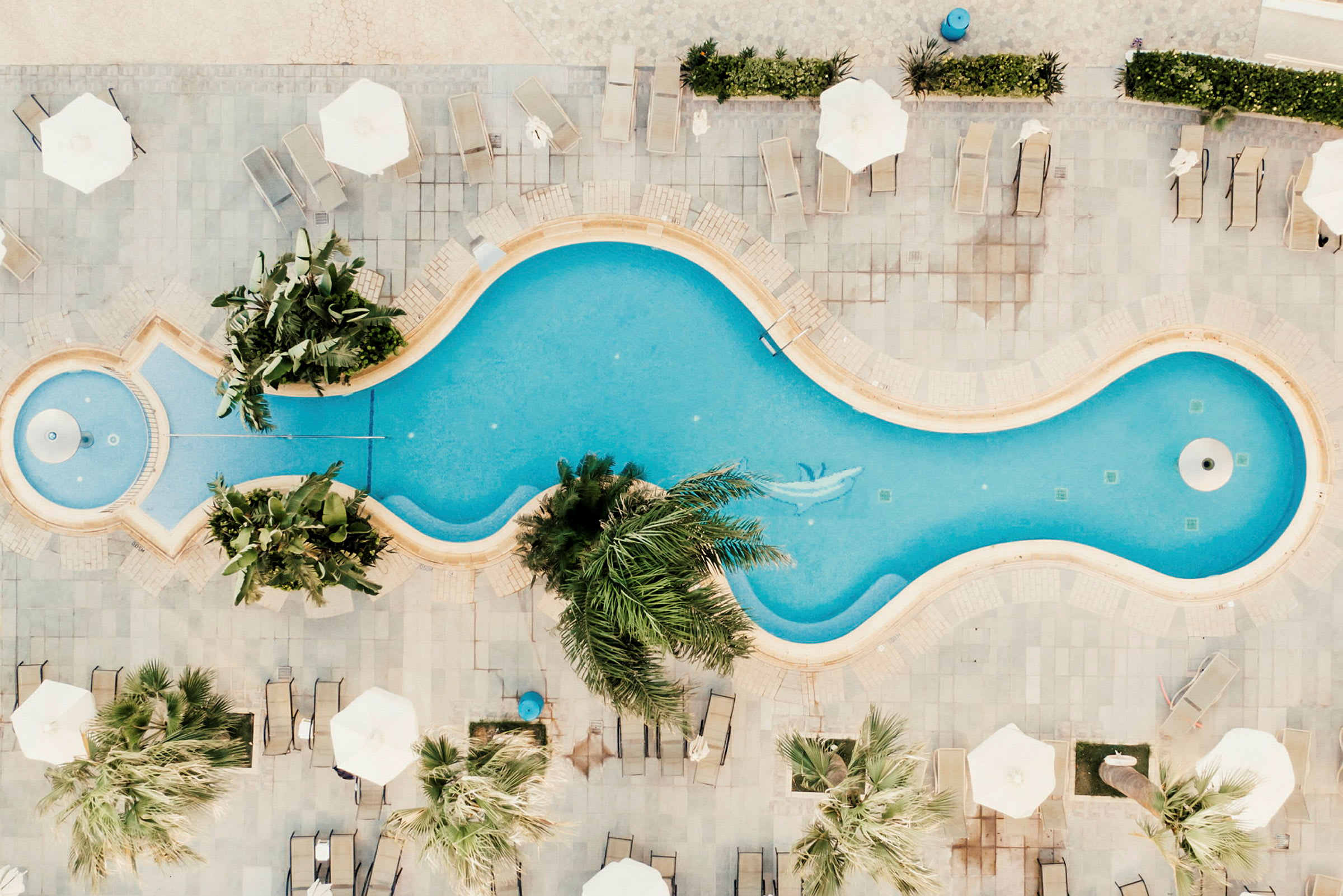 En bild från ovan på en pool med solstolar och parasoller runt omkring samt palmer som omger området. (AI-genererad text)