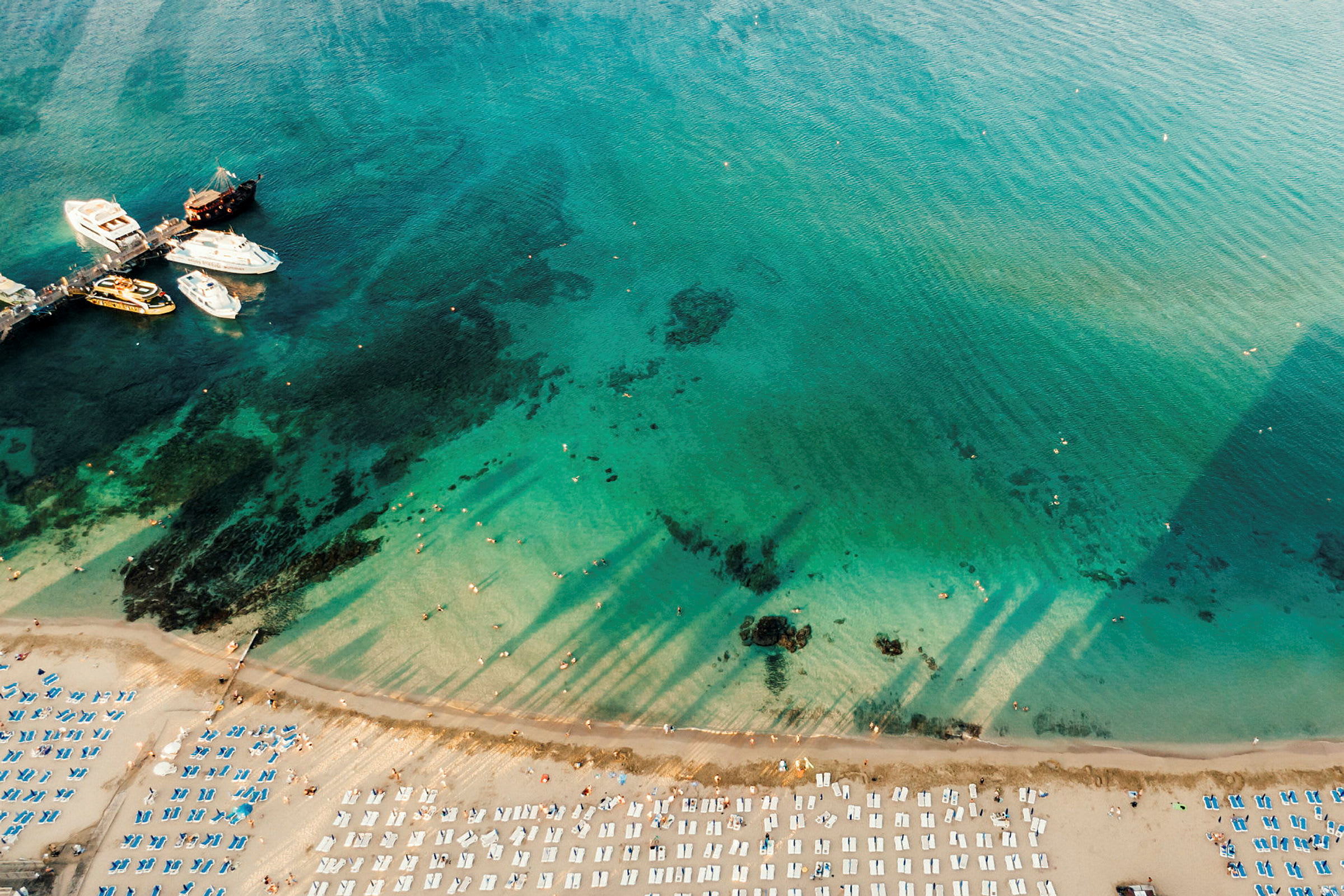 Flygfoto av en strand med solstolar, en brygga och båtar vid havet. (AI-genererad text)