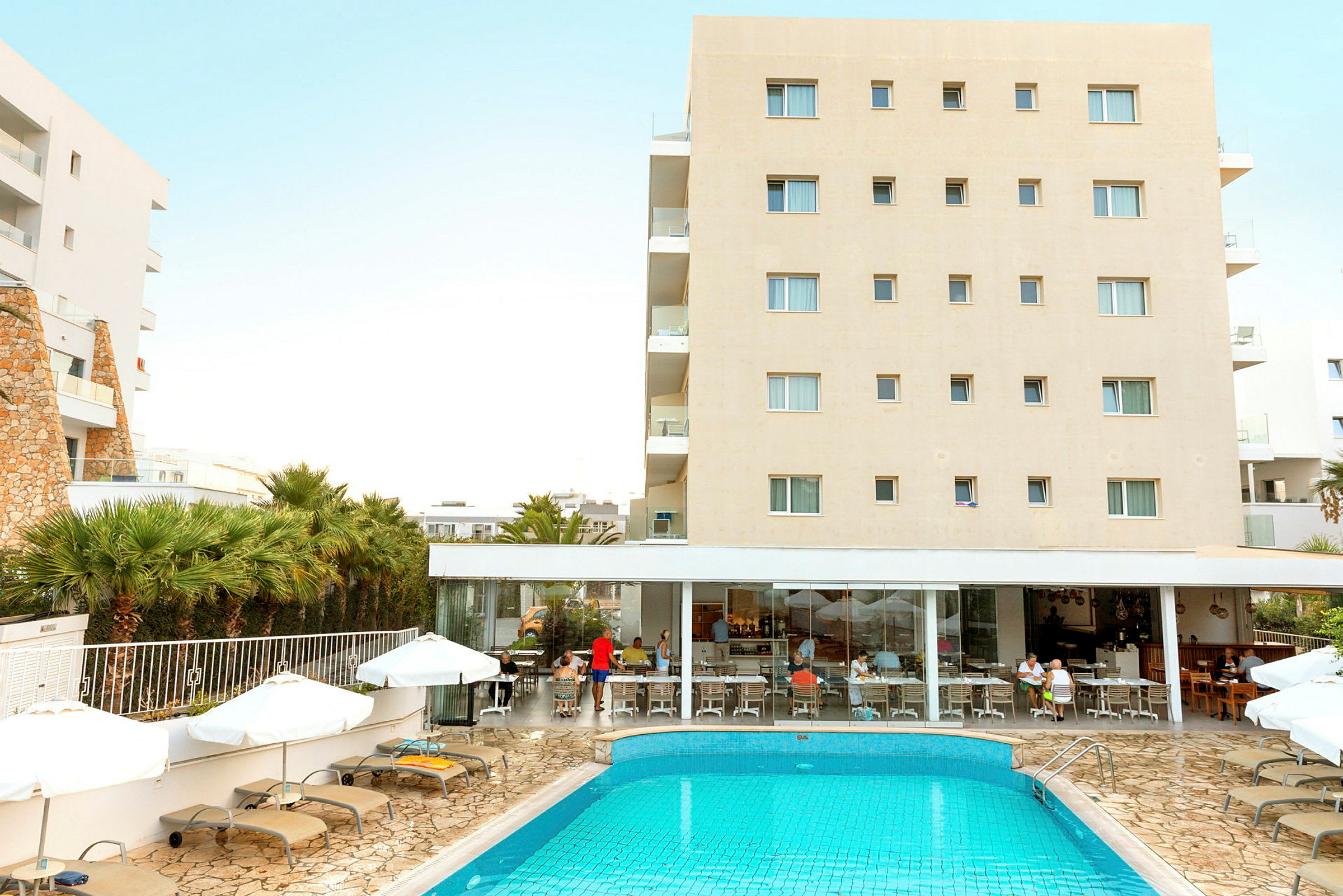 Hotellbyggnad med swimmingpool och solstolar framför. Människor sitter vid restaurangen. (AI-genererad text)