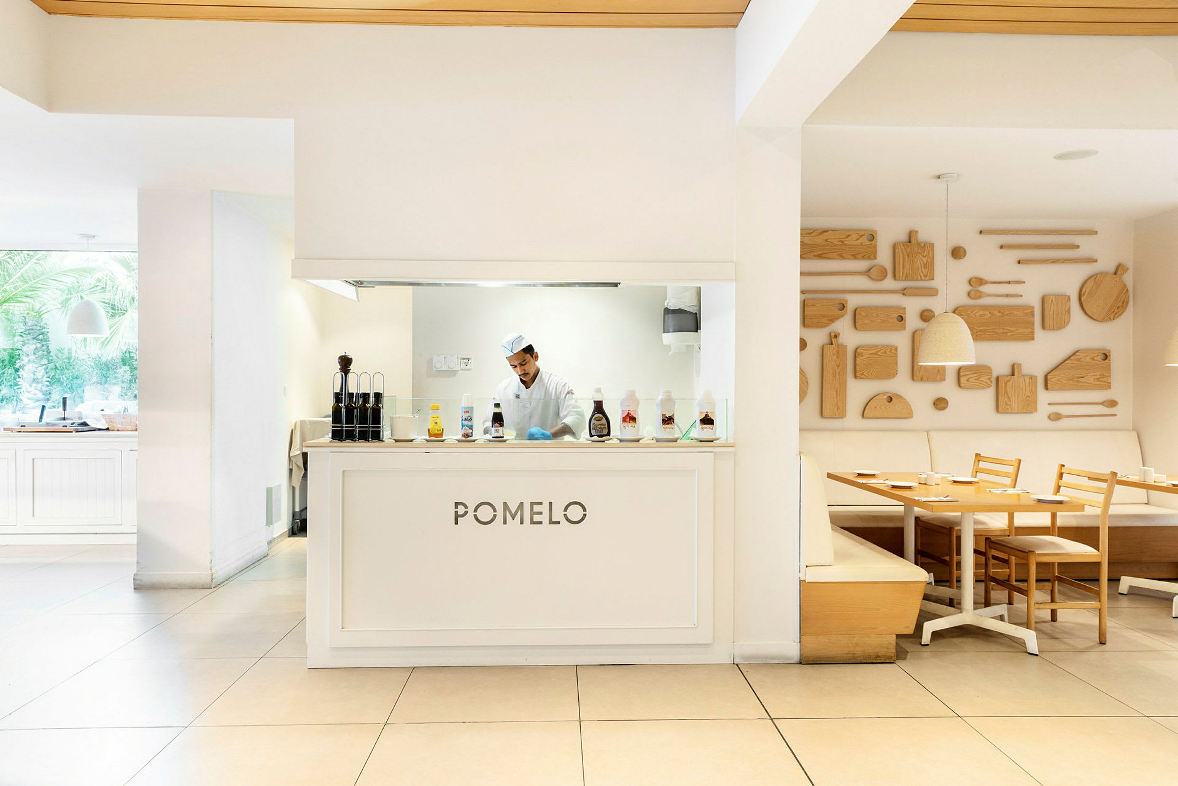 En restaurang med namnet 'Pomelo' och köksområde bakom disken, möblerad med ljusa träbord och stolar samt dekorativa träpaneler på väggen. (AI-genererad text)