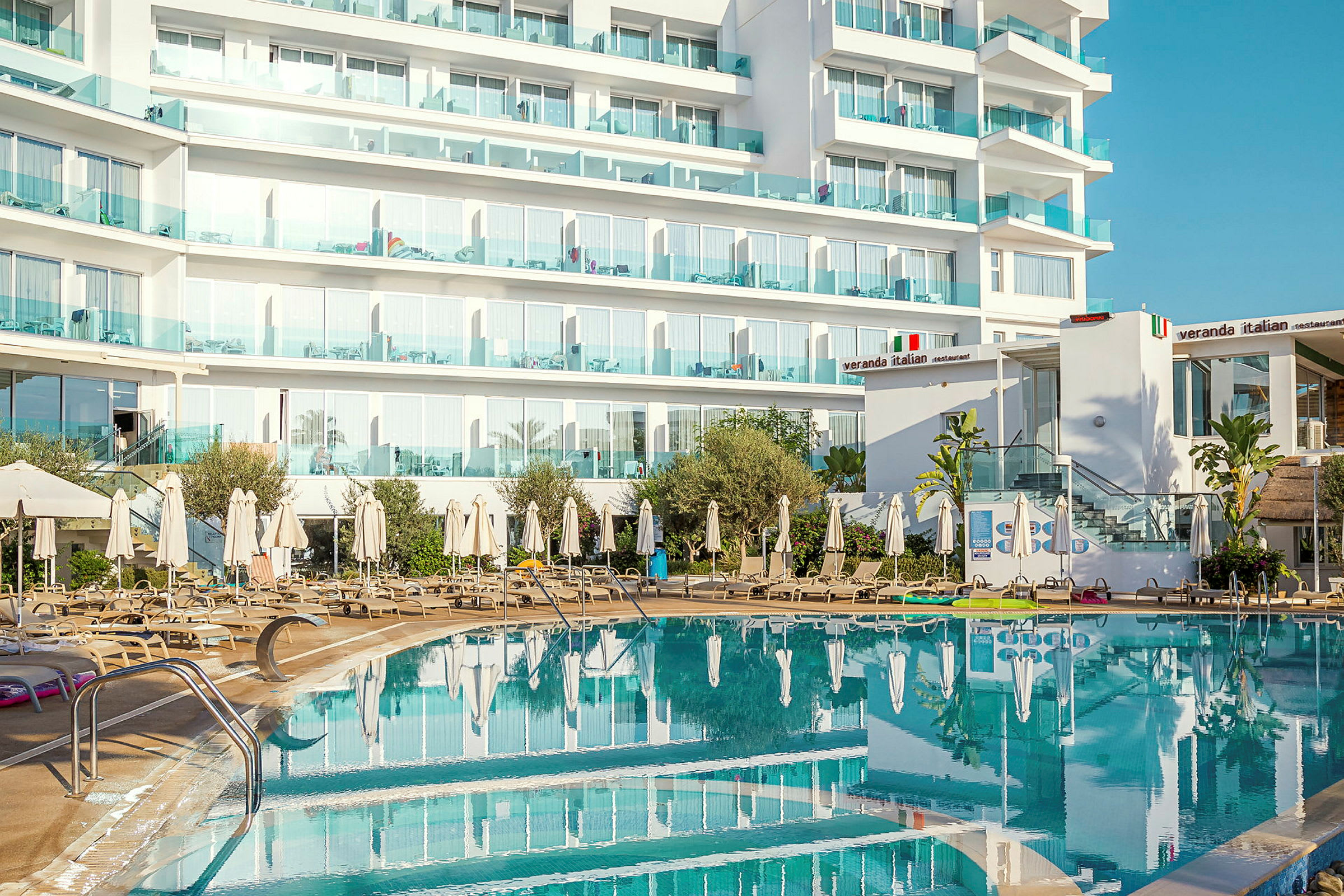 Hotellbyggnad med balkonger bakom en utomhuspool och solstolar. (AI-genererad text)