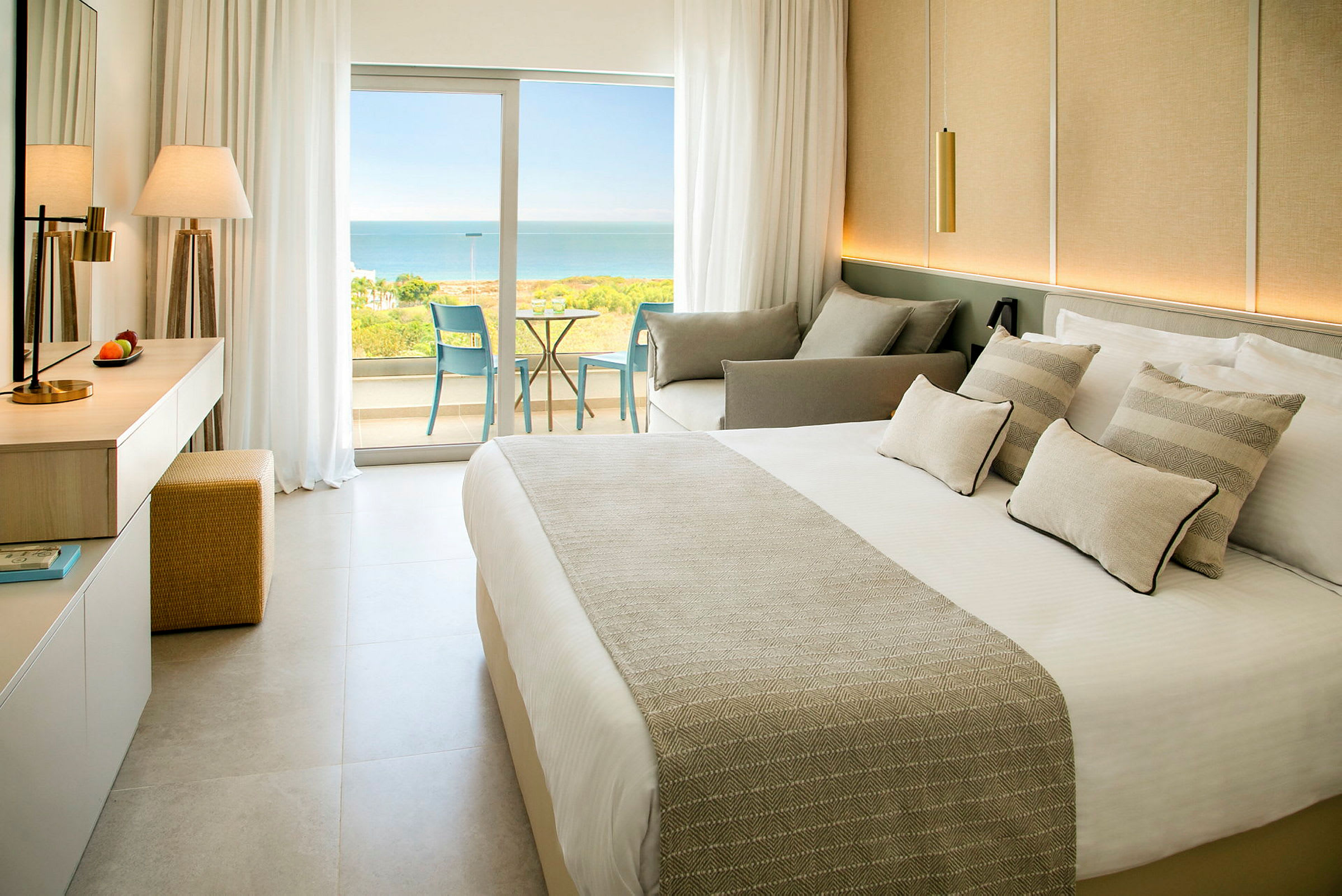 Ett hotellrum med en säng, soffa och glasdörrar som leder ut till en balkong med utsikt över havet. (AI-genererad text)