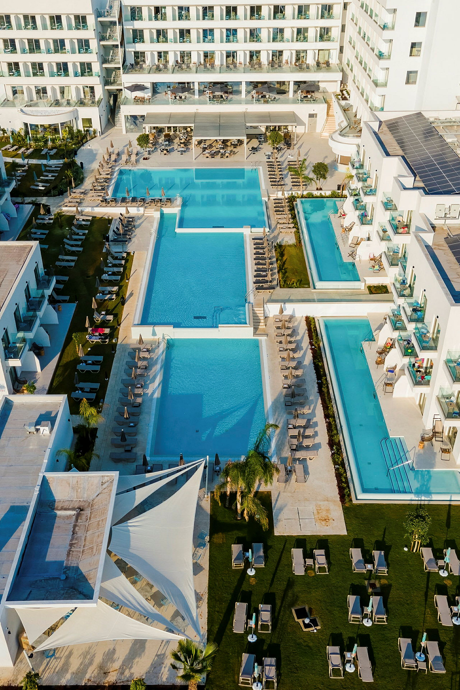 Bilden visar ett hotellkomplex med flera stora poolområden omgivna av solstolar och gröna ytor. (AI-genererad text)